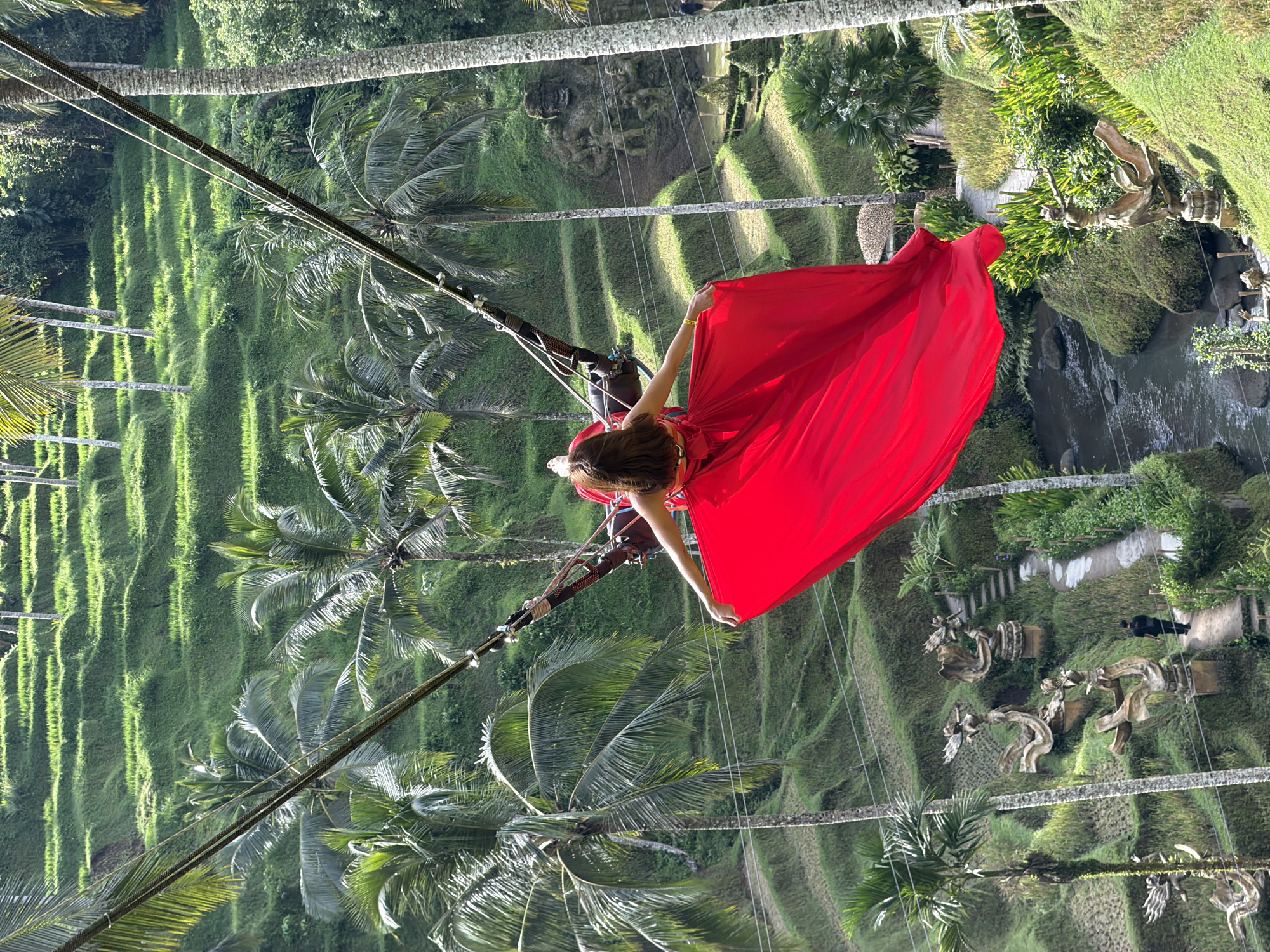 A woman in a red dress is suspended from a swing over the rice terraces of Tegal Suci, Indonesia. The woman's arms are outstretched, holding the dress. The rice terraces are lush and green, with rows of palm trees. A river runs through the valley below, with sculptures and walkways. A person is walking along one of the walkways. The swing appears to be suspended by cables.
