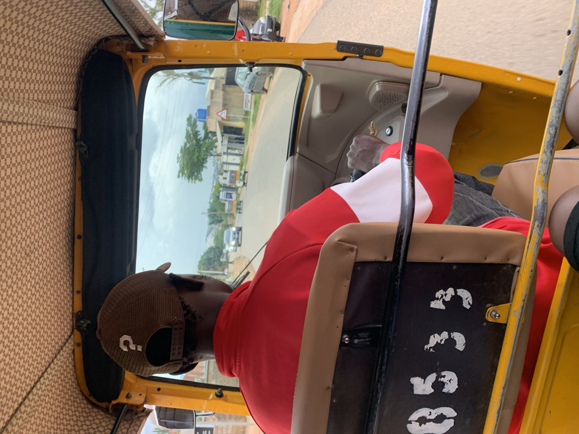 A person wearing a red and white jacket and a brown baseball cap with a white question mark on it is driving a yellow auto-rickshaw. The auto-rickshaw's interior is visible, including a black seat with white stenciled numbers that appear to read "0533". The driver's hands are on the steering wheel. Outside the auto-rickshaw, a road is visible with other vehicles and buildings. The sky is cloudy. The scene appears to be in Gura Top, Nigeria.