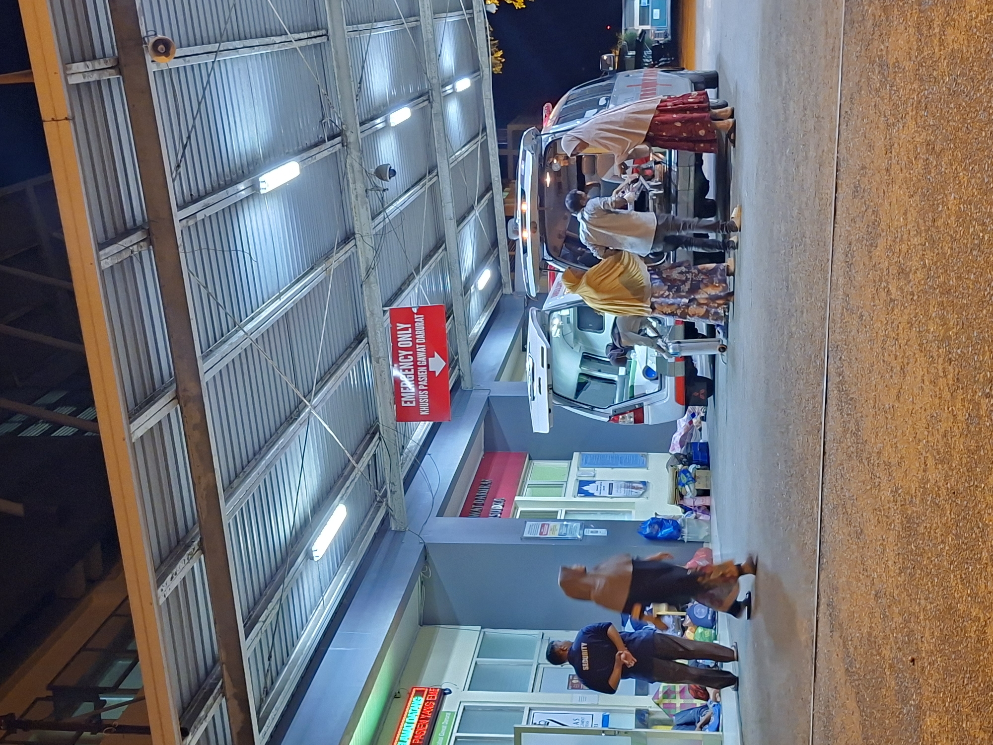 The image captures a nighttime scene at the emergency entrance of a hospital in Banda Aceh, Indonesia. An ambulance is parked under a corrugated metal awning with bright fluorescent lighting. People are attending to someone being transferred from the ambulance, and a security guard stands near the entrance to the hospital. Signage indicates the area is for "Emergency Only" and "Khusus Pasien Gawat Darurat," the latter being Indonesian for "For Emergency Patients Only." Additional signs with Indonesian text are visible. The environment is urban, with a paved surface in the foreground.