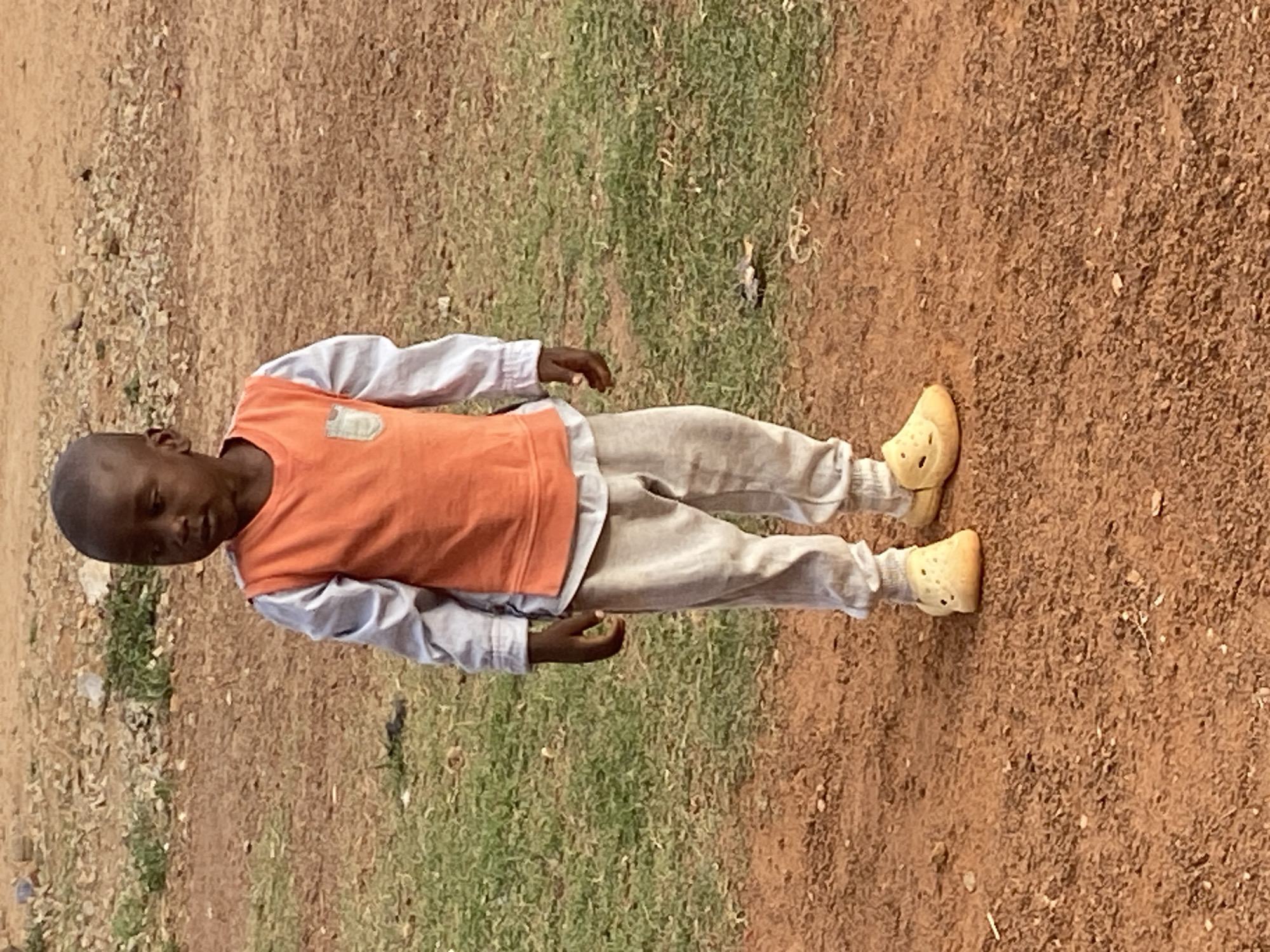 A young child stands outdoors in Bukuru, Nigeria, looking directly towards the viewer. The child, with dark skin and short, dark hair, is dressed in a light-colored (possibly light blue or grey) long-sleeved shirt, over which they wear a vibrant orange sleeveless vest. A light-colored emblem or patch is visible on the upper left side of the orange vest. The child also wears light-colored (beige or light grey) trousers and distinctive light yellow, perforated clogs, resembling Crocs, on their feet.
The setting is an open, natural environment with a ground composed mainly of reddish-brown soil and dirt, interspersed with patches of green and dry grass. Small stones and earth debris are visible on the ground. The lighting indicates it is daytime, with a soft, even illumination suggesting either bright overcast weather or diffused sunlight, as there are no harsh shadows. The child's expression is neutral and observant. There is no visible text identifiable on the child's clothing or within the scene.