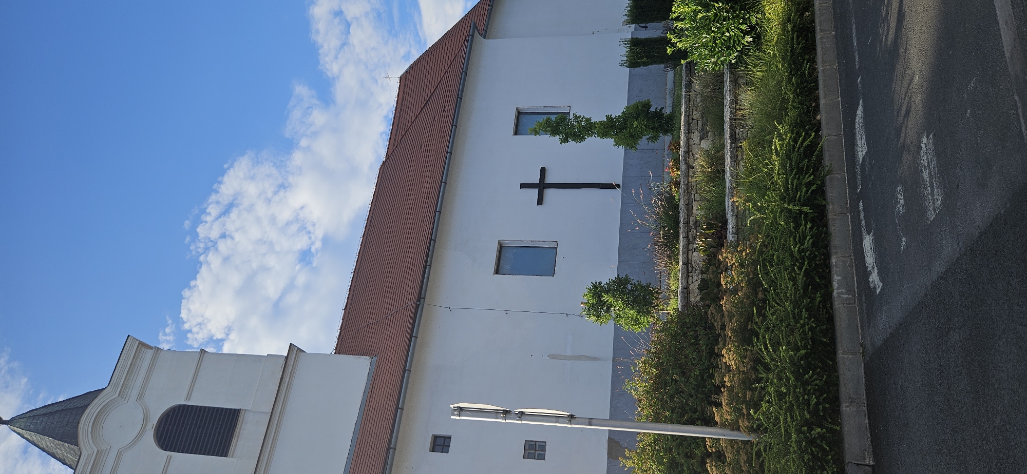 The image depicts the exterior of a white-walled church with a red-tiled roof in Balatongyörök, Hungary. A dark, wooden cross is prominently mounted on the church's facade between two rectangular windows. A white bell tower, partially visible on the left, extends upwards and is topped with a dark spire. The building is situated in front of a bright blue sky dotted with scattered white clouds. In the foreground, a landscaped area features various types of greenery, including bushes and small trees, arranged on tiered stone walls. A grey paved road with white markings is visible at the very bottom of the image. The scene is illuminated by natural daylight.