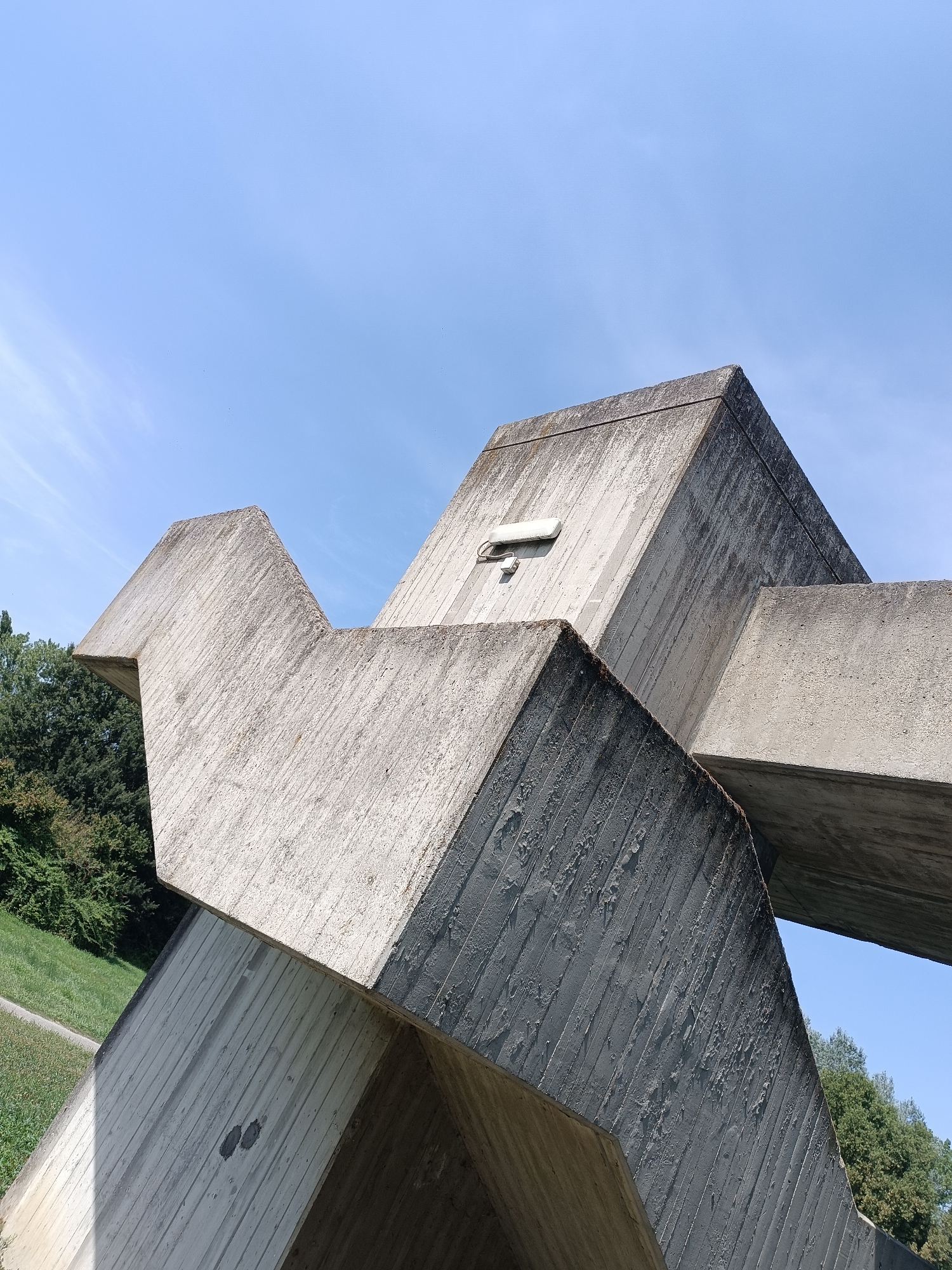 The image displays a close-up, low-angle shot of a large, angular concrete sculpture against a clear blue sky with faint wisps of white clouds. The sculpture, made of rough-textured concrete, is composed of intersecting geometric planes, creating a dynamic and abstract form. The concrete surfaces show vertical striations from the molding process and some weathering, with darker stains and a small, white, rectangular fixture mounted on one of the upper surfaces. To the left and background, a grassy slope with some trees is visible, suggesting an outdoor setting in a park or open area. The scene is bathed in bright daylight, casting subtle shadows that emphasize the sculpture's sharp edges and volume. The location is identified as Casalmaggiore, Italy.