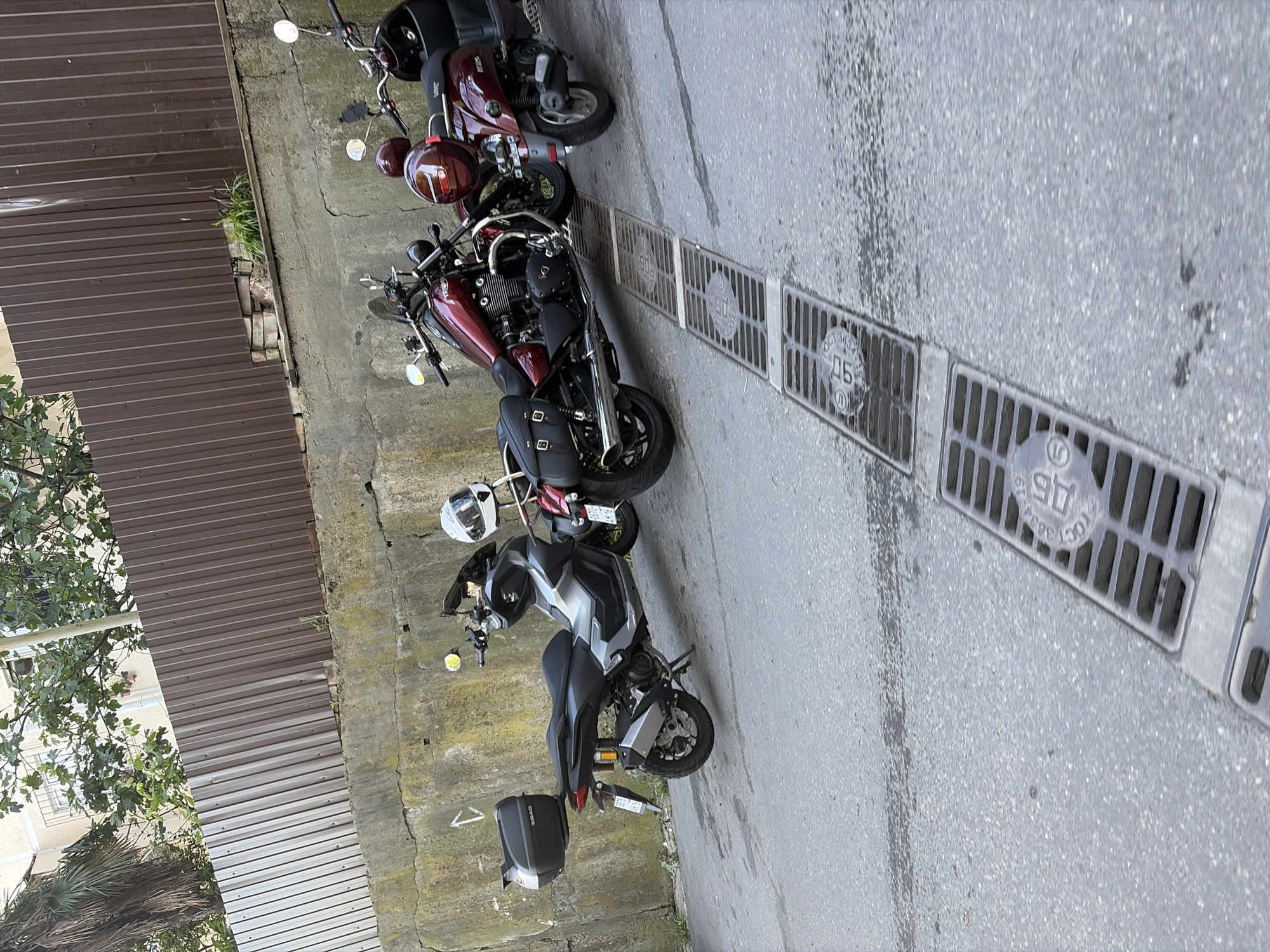 This outdoor image, taken in Sochi, Russia, shows several motorcycles and scooters parked parallel to a wall and a corrugated metal fence on a sunny day. The scene depicts a side road or alleyway with asphalt pavement featuring a row of rectangular metal drainage grates.
The subjects include a grey and silver modern scooter or small motorcycle with a GIVI S340 top case and a white helmet resting on its seat. Next to it is a dark red cruiser-style motorcycle equipped with black saddlebags. Further along are two more scooters, one dark red with a matching helmet, and another partially visible in the background.
No explicit activity is taking place, but the vehicles are parked, suggesting a break or storage.
Notable details confirm the Russian location: the drainage grates prominently display Cyrillic text, including "ГОСТ 3634-19" (GOST 3634-19, referring to a Russian State Standard) and the letters "ДБ" (DB), along with numbers like "90" and "96". The bright daylight and clear conditions indicate good weather. Lush green foliage, including palm trees and leafy trees, is visible, consistent with Sochi's subtropical climate. A concrete wall runs alongside the road, behind which stands a tall brown corrugated metal fence. Buildings are faintly visible through the trees in the background.