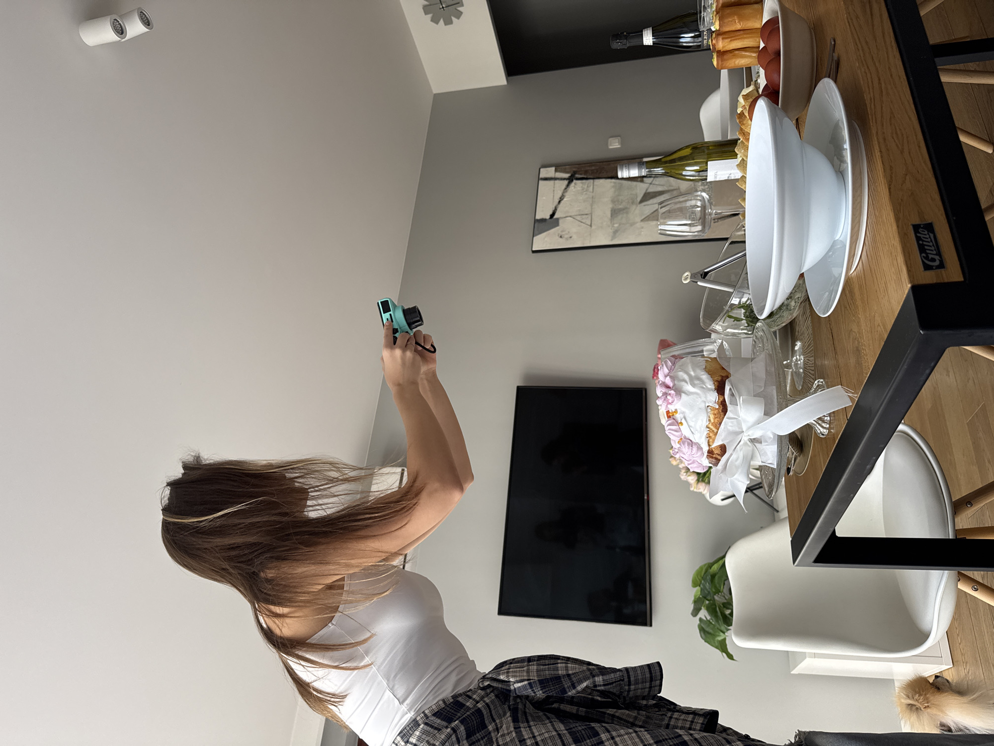 A young woman with long brown hair, wearing a white tank top and a plaid shirt tied around her waist, is holding a small, blue camera and taking a picture. The scene is likely inside a modern apartment. In the background is a dining table set with food items, including a cake decorated with pink and white flowers and a white bow, along with plates, bowls, glasses, and bottles of wine. The table has a light-colored wooden surface with a black metal frame. Near the table, there is a white chair and a small dog lying on the floor. On the wall is a television and an abstract art piece. Based on these details, the location could be a residential apartment in Warsaw, Poland.