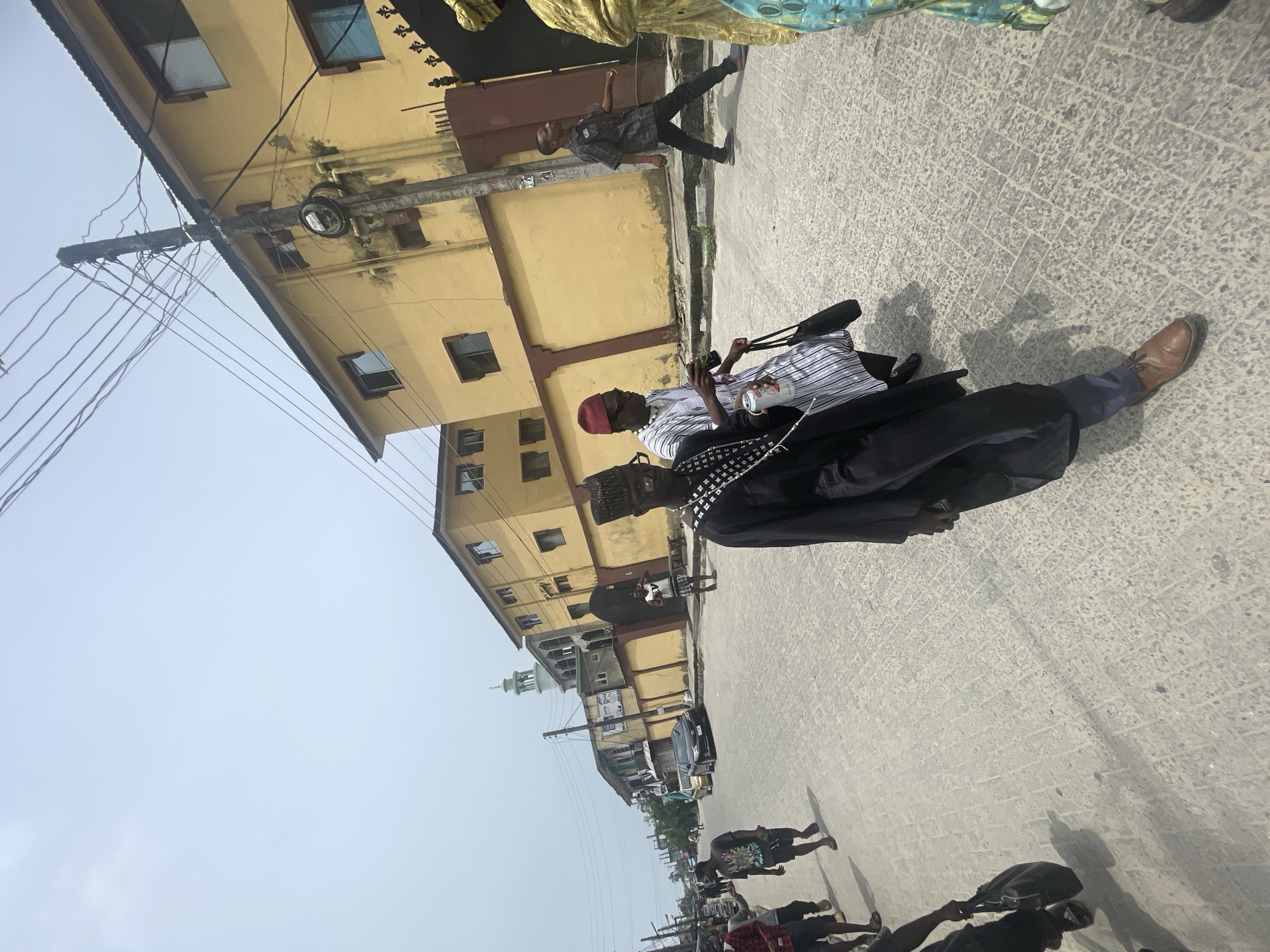The image captures a street scene in Ado, Nigeria. The environment features multi-story yellow buildings, power lines, and a paved street. Several people are present, including a man with a red cap and a black jacket, seen holding what appears to be a thermos. People walking in various directions are visible in the street. A green-domed minaret from a mosque stands in the background. Vehicles are parked along the street.