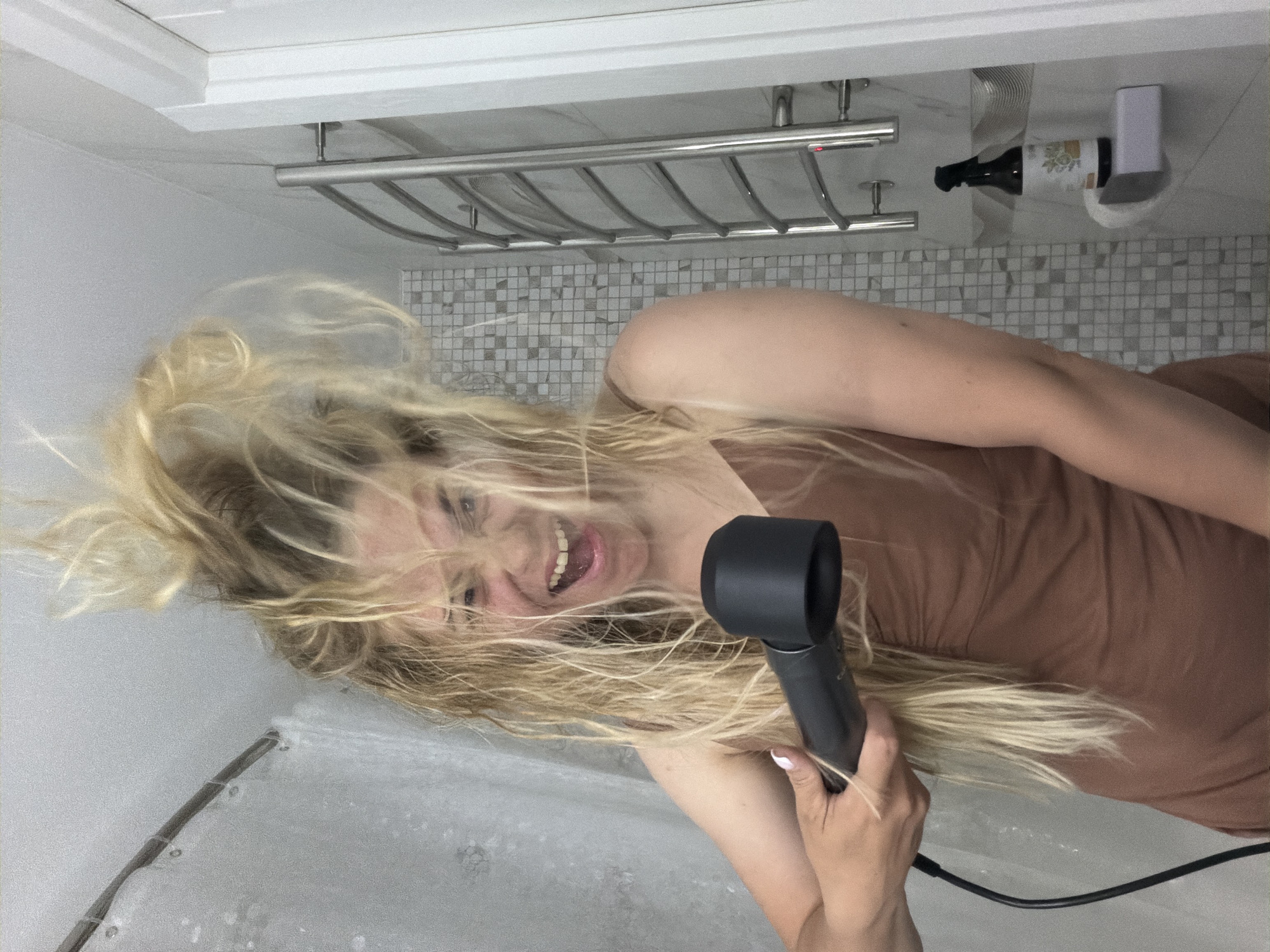 A woman with blonde hair is holding a black hairdryer and smiling in a bathroom. The bathroom has a chrome towel rack, gray mosaic tile walls, and a shower with a glass door. A bottle of liquid is on a shelf in the corner. The exact location in the city of Votkinsk, Russia is not discernible from the image.