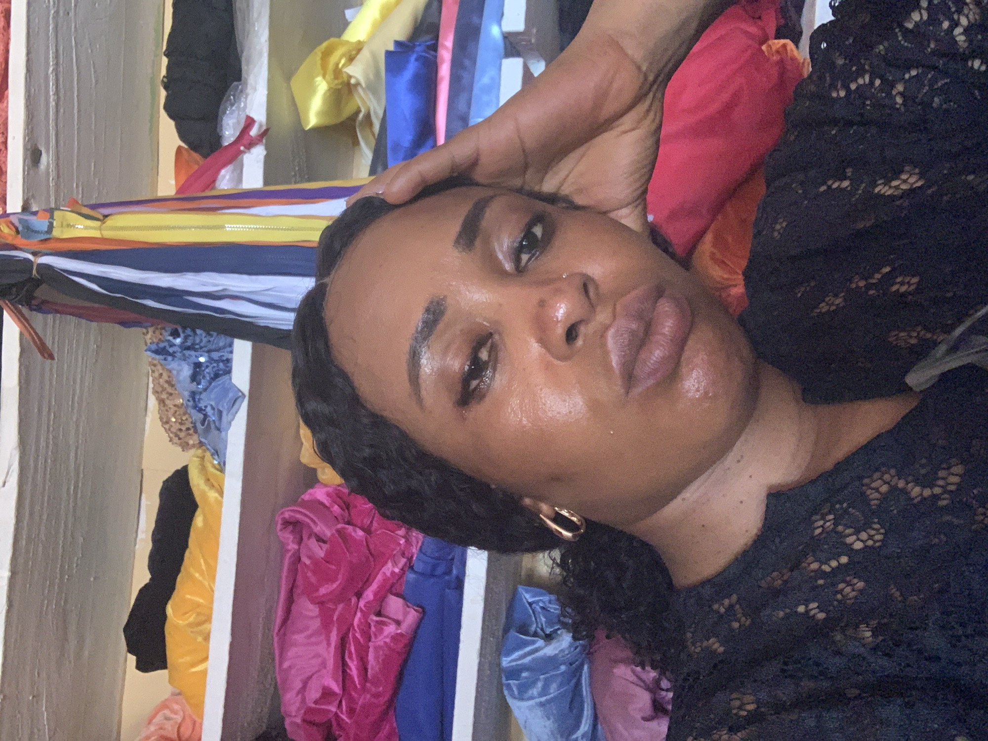 A medium, close-up shot of a dark-skinned person with black, curly hair wearing a black, lace top. The person is lying down with their hand on their forehead, facing the camera. Their skin appears glossy, possibly due to sweat or moisturizer. Behind them, there is a shelf containing folded pieces of colorful fabrics, including yellow, blue, pink and red. This image was taken indoors. There is no evidence to suggest the location of Sabon Tasha, Nigeria.