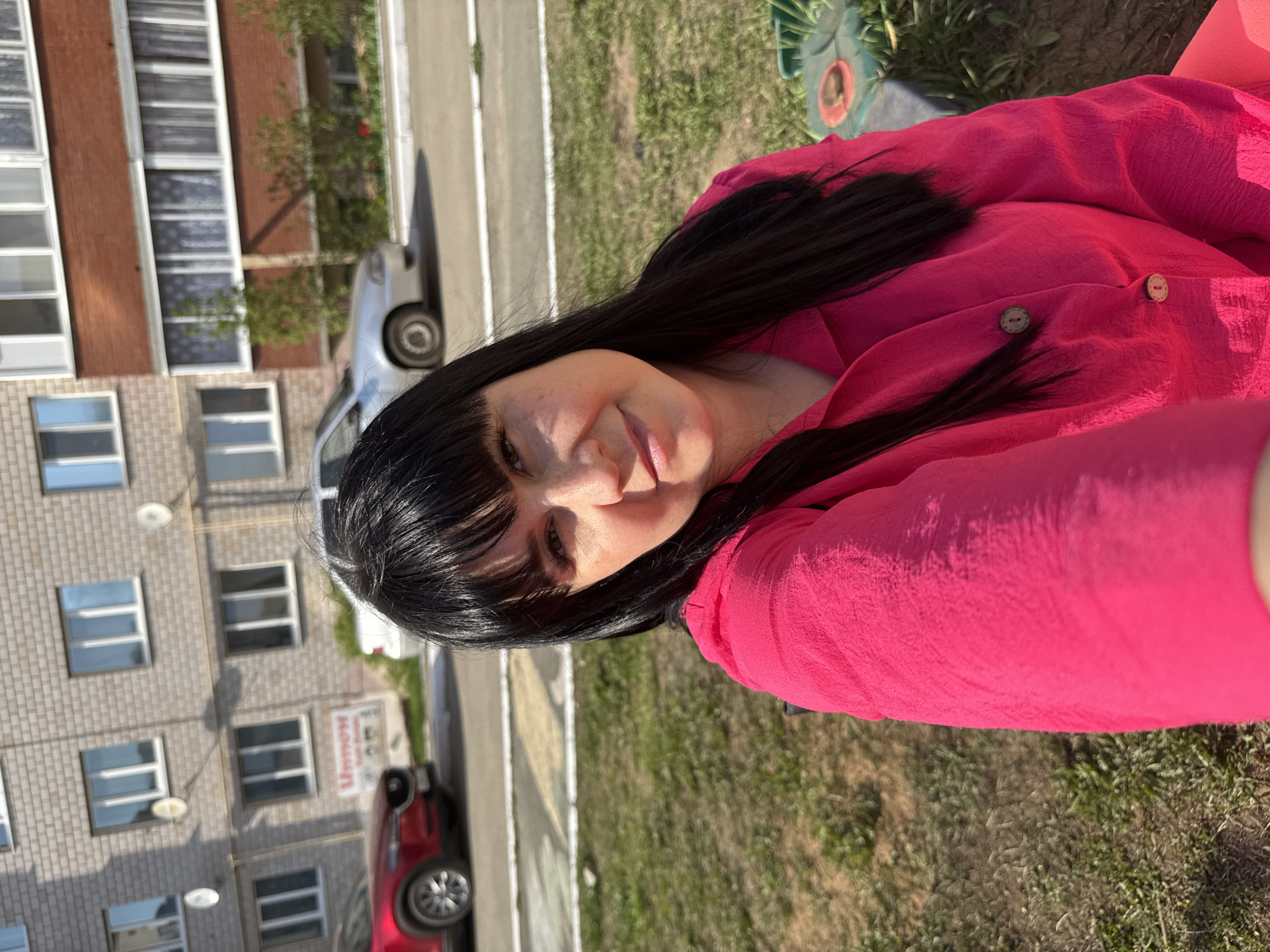 A woman with long, dark hair and bangs is wearing a bright pink button-down shirt and is positioned in an outdoor setting. She appears to be on grass, with part of a green and gray object possibly a piece of playground equipment visible behind her. In the background, there's a multi-story building with numerous windows. A street runs along the side of the building, and cars are parked along the curb. Based on the provided location, this scene takes place in Zav'yalovo, Russia. The woman is looking towards the camera with a slight smile.