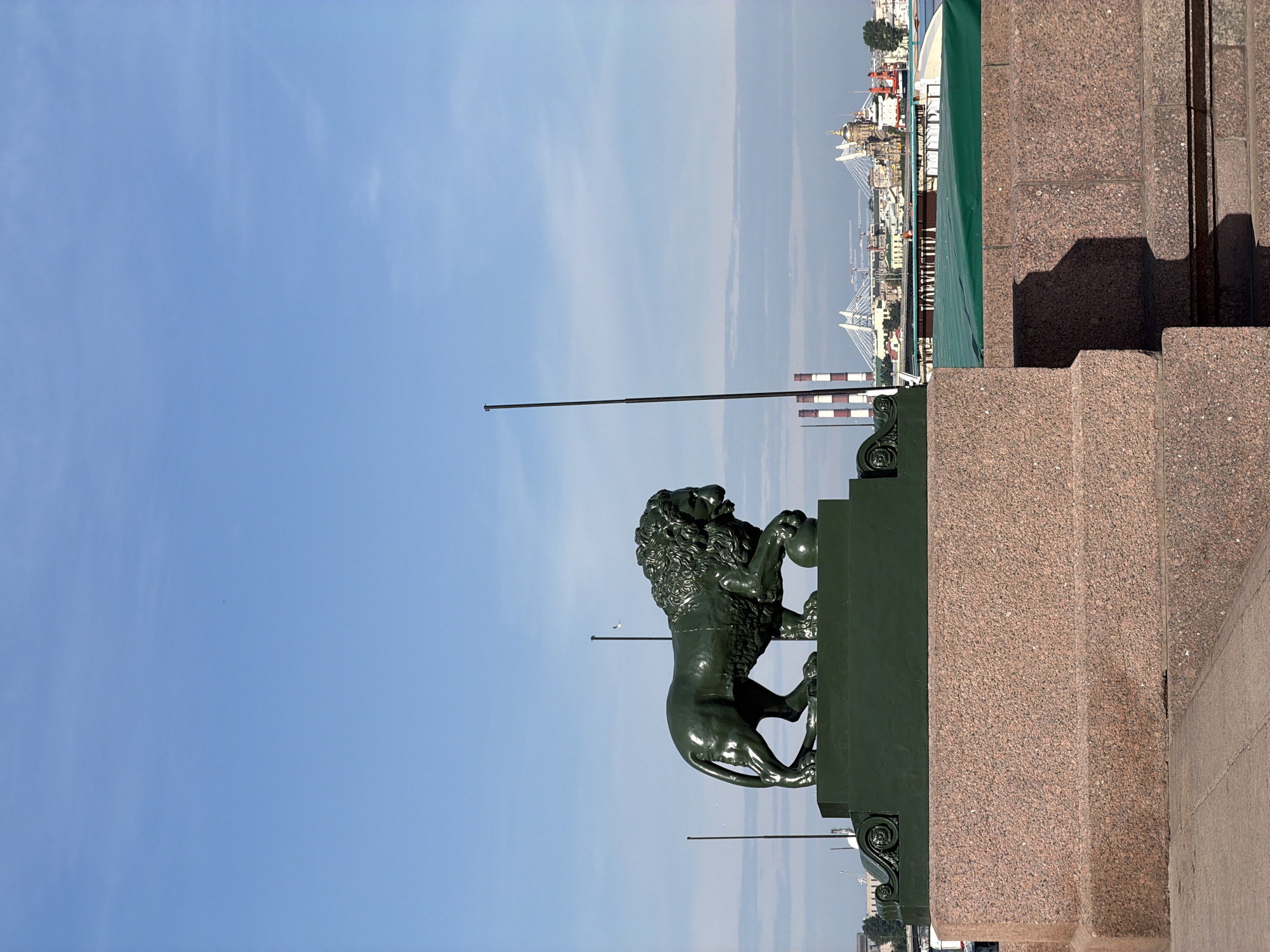 This is an outdoor, eye-level, medium shot photograph taken on a sunny day in Saint Petersburg, Russia. The primary subject is a dark green bronze statue of a lion, standing on its hind legs with its front paw resting on a sphere, which is mounted on a pedestal. The statue is positioned on the left side of the frame, with its head facing to the right. Behind the statue, the sky is a clear, pale blue with some wispy white clouds. In the background, there is a cityscape with buildings, a bridge with white cable-stays, industrial smokestacks, and a ship. The foreground consists of a textured granite structure, possibly a bridge or embankment, in shades of pink and gray. The scene depicts an urban environment with a notable monument against a backdrop of city infrastructure and natural elements.