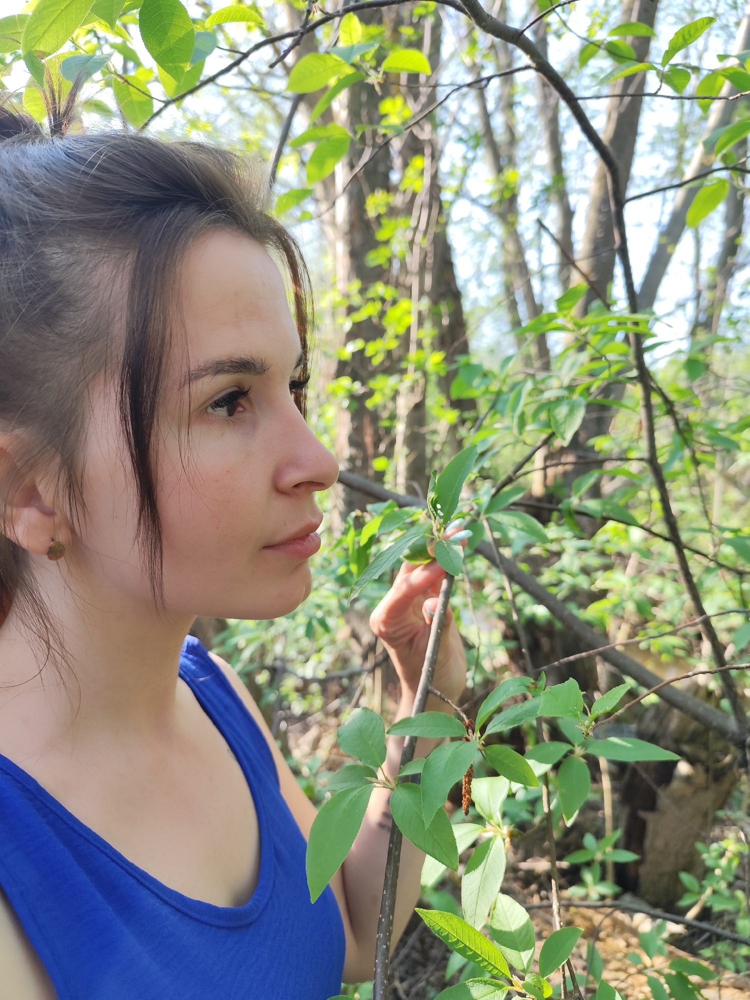 A woman with brown hair pulled back in a bun, wearing a blue tank top, stands outdoors in what appears to be a wooded area. She is looking to the right, possibly examining a branch with green leaves she is holding. The location is likely Фоки, Russia, given the context. The environment consists of trees and greenery.