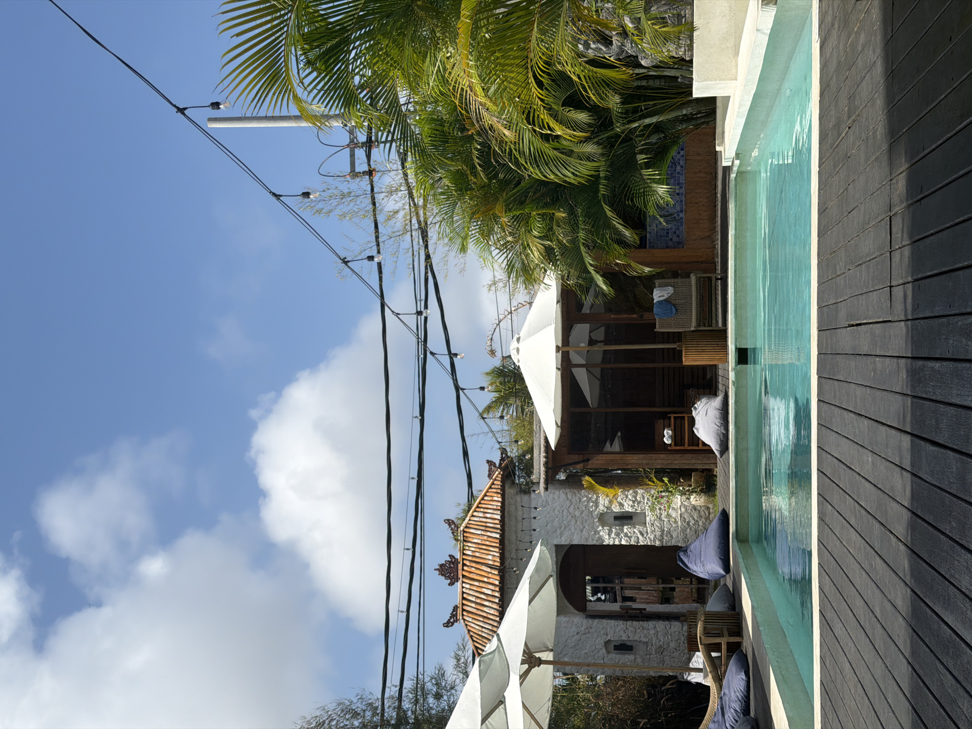 A wide shot captures an outdoor swimming pool area, likely in Kuta Selatan, Indonesia, on a bright, cloudy day. The foreground is dominated by a dark wooden deck that leads to a rectangular swimming pool filled with clear turquoise water. Along the left edge of the pool, several blue and gray beanbag chairs are arranged on the deck. To the left of the pool is a white stone building with an arched entryway and a traditional tiled roof featuring ornate detailing. A white umbrella stands to the left of the building. To the right of the pool, there is a wooden structure with an open-air section covered by a white umbrella, and a single lounge chair with towels draped over it. Beyond this, lush green palm trees and tropical foliage fill the right side of the frame. A string of bare lightbulbs is suspended across the top of the image, strung between poles and trees, suggesting the area might be used at night. The sky above is a vibrant blue, interspersed with fluffy white clouds. The scene evokes a relaxed, tropical resort atmosphere.