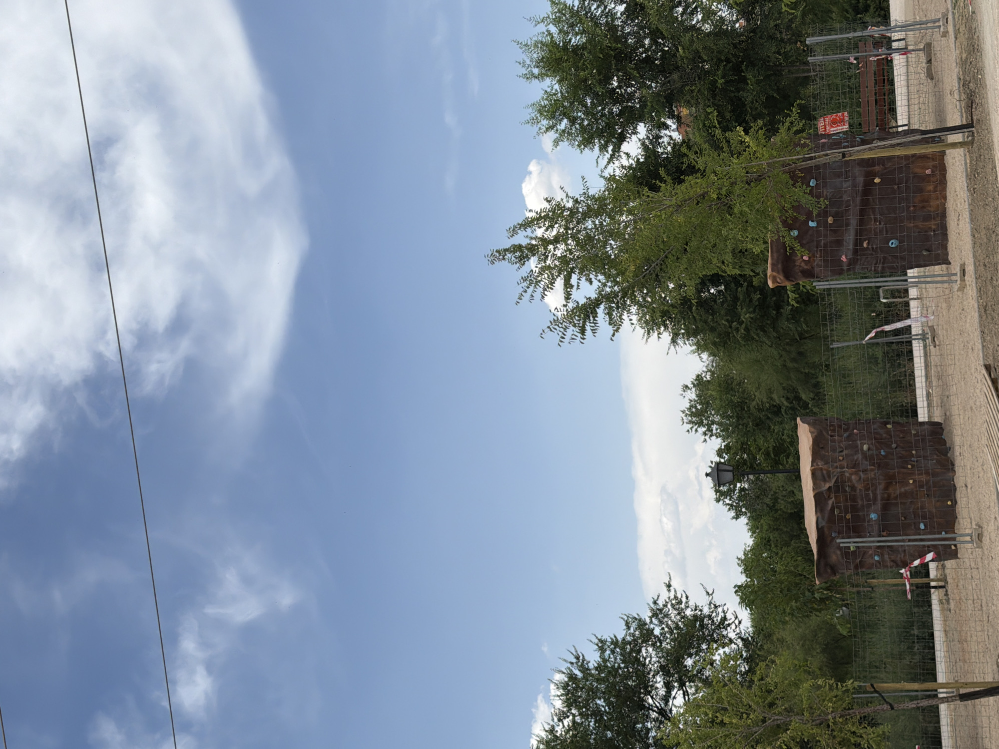 This is a vertical-format outdoor photograph taken on a sunny day with a clear blue sky and scattered white clouds. The image captures a segment of what appears to be a park or recreational area.
The primary subjects in the foreground are two artificial climbing walls. These walls are textured and brown, with colorful climbing holds attached. They are enclosed by wire fencing, with a few red and white caution tapes visible. Behind the climbing walls, dense green trees form a backdrop, some of which overhang the scene. A black lamppost is also visible near one of the climbing walls.
In the distance, more trees and foliage suggest a natural environment. The ground appears to be a sandy or gravelly pathway. A red and white sign with some text is affixed to the fencing of one of the climbing walls. The overall impression is of a sunny, pleasant day at an outdoor activity venue. The image is rotated, suggesting it was taken with the phone in a portrait orientation.
