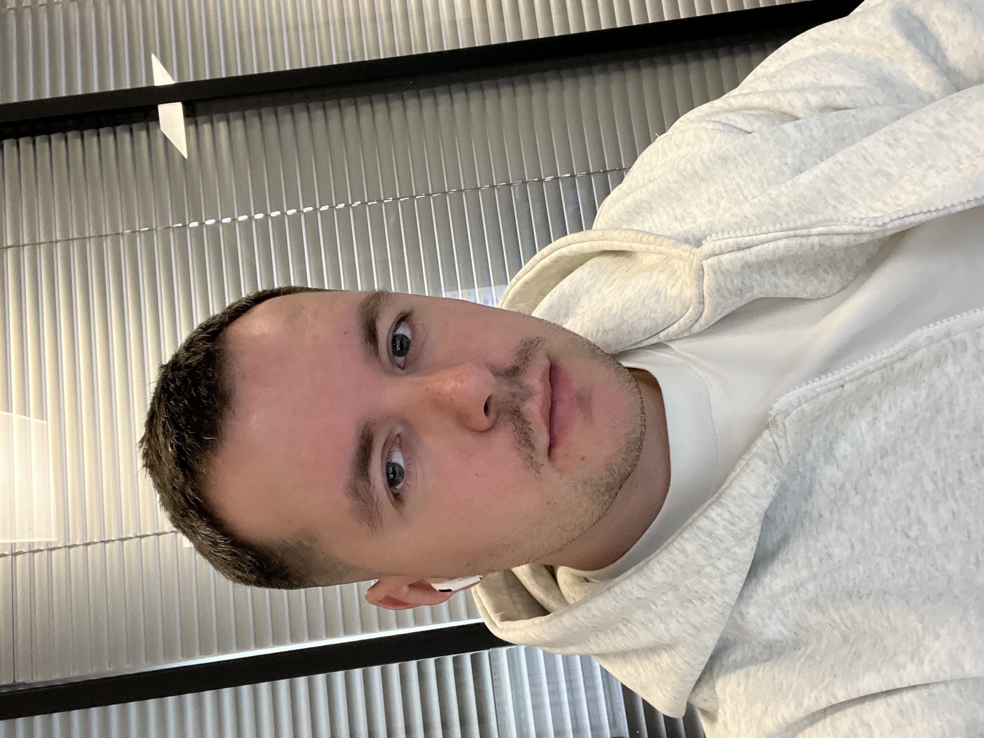 A portrait-style image, rotated 90 degrees clockwise, prominently features a young man from the chest up. He has short, dark hair with a faded cut on the sides, light skin, and light stubble on his chin and upper lip. His eyes, a shade of blue or green, gaze directly towards the viewer with a neutral expression. He is wearing a light grey, subtly textured hooded sweatshirt over a white t-shirt. A white wireless earbud is visible in his right ear. The background is composed of a series of light-colored, vertical ridged panels or blinds, with a dark horizontal bar crossing the upper part of the frame. The lighting is soft and even, suggesting an indoor setting. No text is visible in the image. The scene is located in Moscow, Russia.