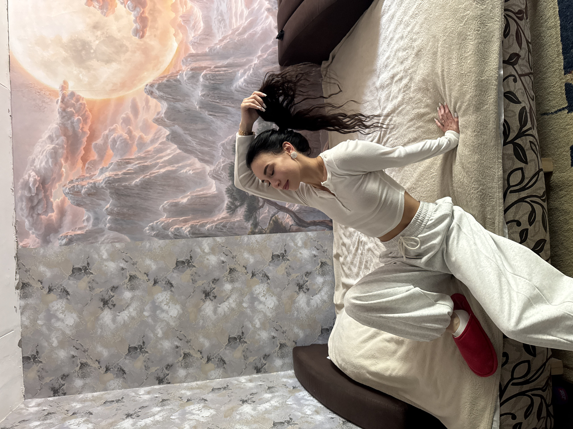 A woman is lying on a bed. She is wearing a white long-sleeved top and gray sweatpants. She has one red slipper on her foot. She is holding her black hair up in a ponytail. The bed has a cream-colored patterned bedspread and a brown headboard. The wall behind the bed is painted with a mural of a moon and clouds. The lower part of the wall has a gray and white marble-like pattern. The location is in Chaikovsky, Russia.