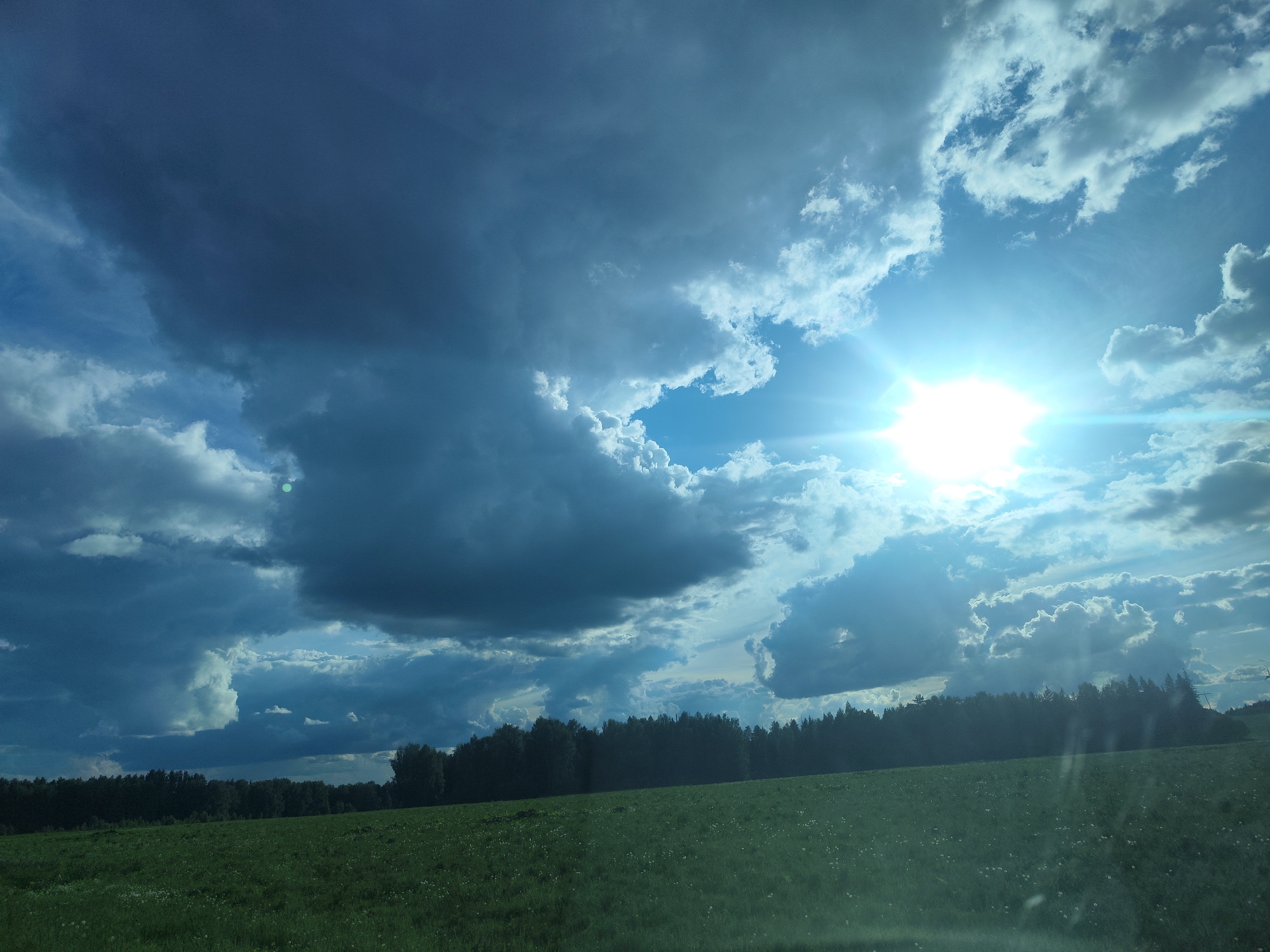 The image shows a landscape with a green field in the foreground, a dark line of trees in the midground, and a cloudy sky in the background. The sun is visible through a gap in the clouds, creating a bright glare. The location is most likely near Альняш, Russia, based on the prompt. The scene depicts a rural environment with a mix of open land and forested areas.