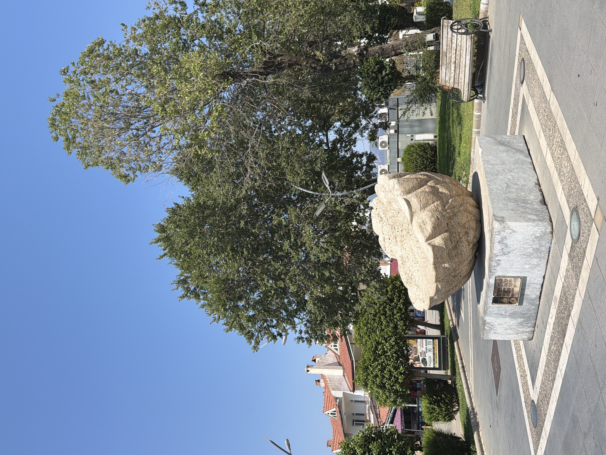 The photograph captures a public square in Marmaris, Turkey, on a clear, sunny day. Dominating the foreground is a large, abstract stone sculpture resembling a cracked shell, resting on a granite pedestal. A plaque is affixed to the pedestal. In the middle ground, lush green trees flank the sculpture, with one particularly large tree to the right. Behind the trees, a row of buildings with red-tiled roofs and white facades are visible, indicative of Mediterranean architecture. Several air conditioning units are mounted on one of the buildings. A vintage wooden park bench with decorative metalwork sits to the right of the sculpture, positioned on a paved pathway. The ground is paved with grey tiles and features a decorative border of smaller grey stones and white lines. The overall environment suggests a well-maintained public space, likely a park or plaza, enjoyed by locals and tourists alike. No people are present in the frame.