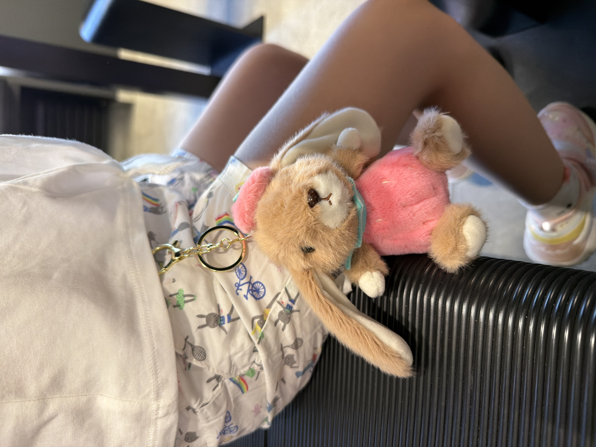 A person is lying down with a plush bunny toy. The person is wearing shorts with a pattern of bicycles, tennis rackets, and figures, along with pink and white sneakers. The bunny toy has light brown fur, pink ears and body, and a light blue ribbon around its neck. The person's clothing is connected to the bunny toy with a gold-colored chain with a ring. The person is lying on a corrugated, possibly black, surface, likely a suitcase or other piece of luggage. The location is unknown and cannot be determined to be Izhevsk, Russia based solely on this image.