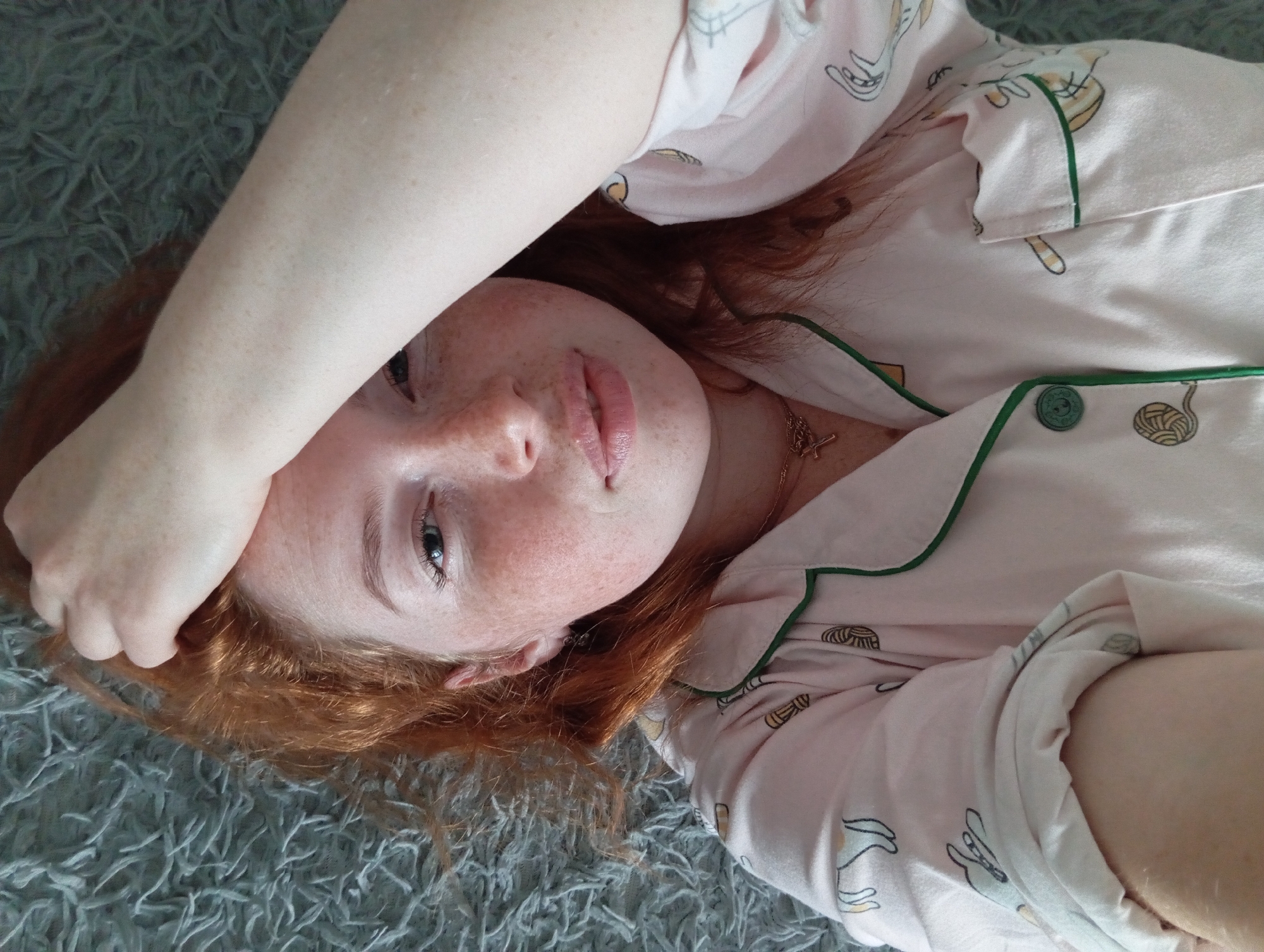 A fair-skinned woman with freckles and red hair lies on a gray shag rug. She is wearing a light pink pajama top with green trim and cartoon cat and yarn ball designs. Her right arm is raised, with her hand near her forehead. She is looking directly at the camera. A gold necklace is visible around her neck. The location is not evident in the photograph.