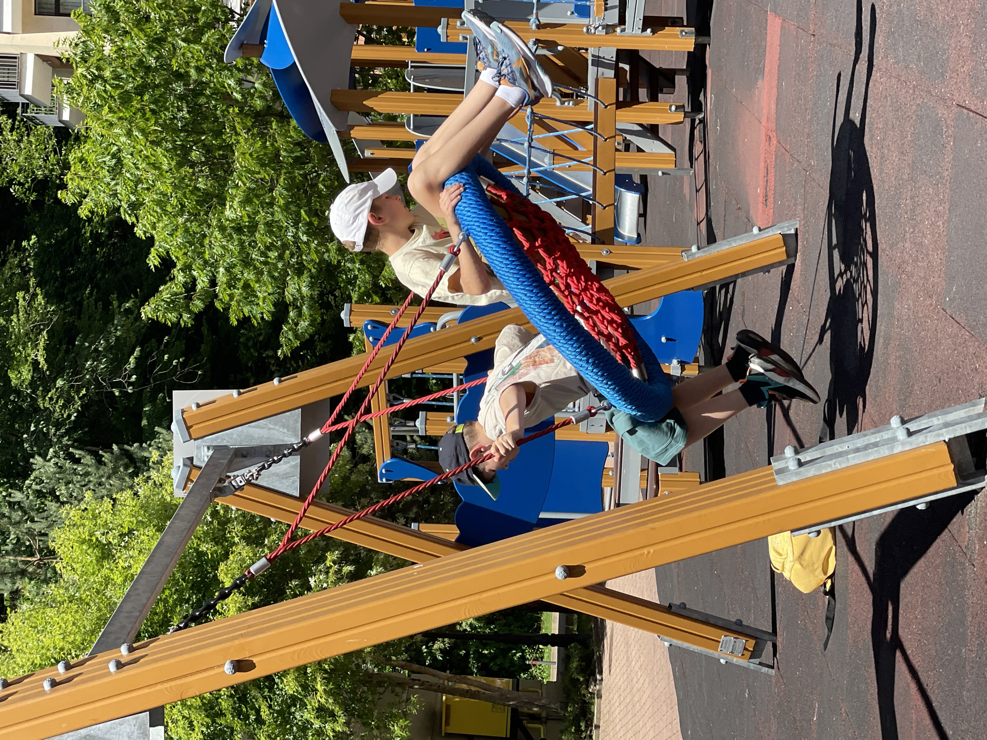 Two young boys are playing on a playground in Estosadok, Russia, on a sunny day. The boys are on a swing set, with one boy sitting on a large, blue and red rope swing. The other boy is standing on the swing, with his arms extended. The playground equipment is made of wood and metal, with a rubberized surface on the ground. There are trees and buildings in the background.