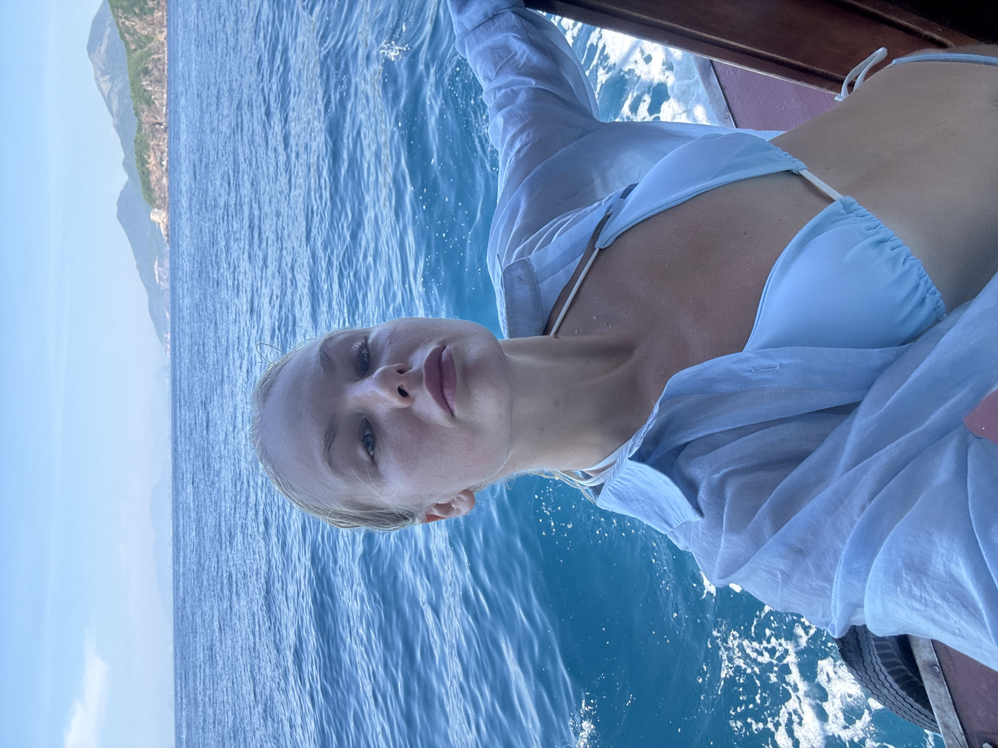 A young woman with blonde hair, wearing a light blue bikini and a sheer light blue button-up shirt, takes a selfie on a boat in the ocean. She is positioned on the right side of the frame, with her face turned towards the camera and her lips slightly parted. Her wet hair is pulled back. The clear blue water of the ocean fills the background, with distant green hills and mountains visible on the horizon. The boat's wooden railing and a tire are visible on the right and left sides of the frame, respectively. The image appears to be taken in Nha Trang, Vietnam, likely in the waters surrounding the city, characterized by its coastline and nearby islands.