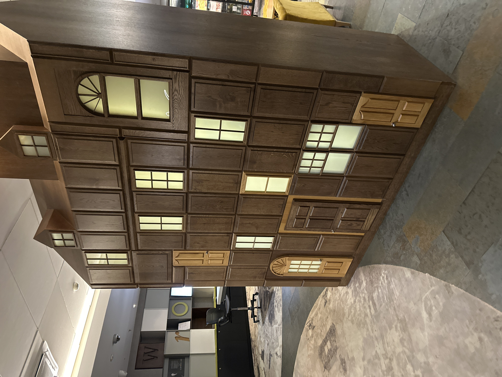 The image shows an interior setting, likely a commercial space, featuring a large, house-shaped wooden structure. The structure is intricately designed with various window and door-like panels, some of which are illuminated. The design is reminiscent of a whimsical, storybook architecture.
In the background, there is a modern office area with desks, chairs, and shelving. The flooring appears to be a combination of tile and textured carpeting. The lighting is predominantly artificial.
Given the specific architectural style and ambiance, the image is likely of a unique feature within a commercial or public space.
The location within Wrocław, Poland cannot be determined with certainty from the image alone. However, assuming it is a public commercial space, the image likely comes from a public structure somewhere in Wrocław, Poland.