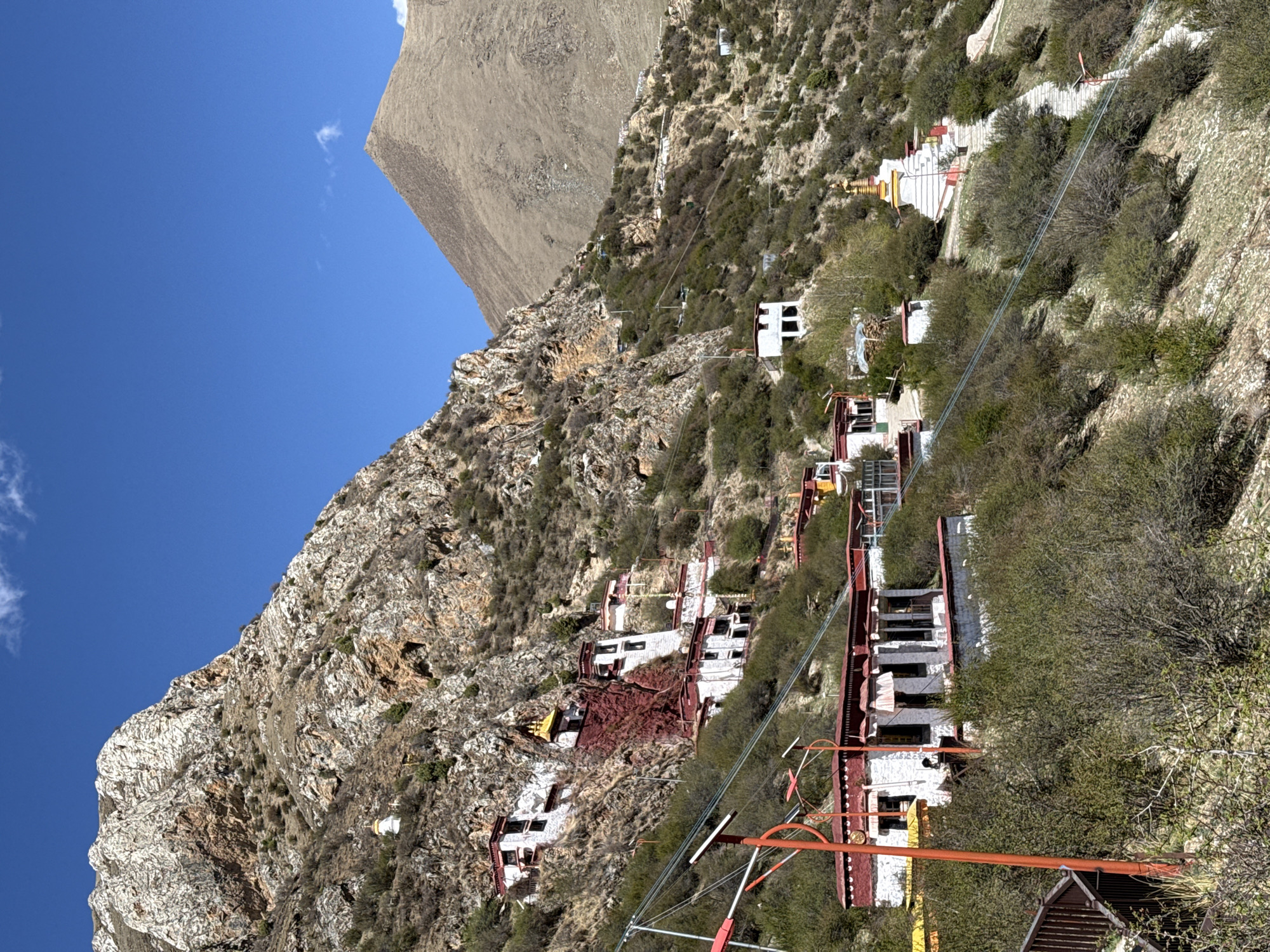 The image shows a mountain slope with buildings and a cable car system. The buildings appear to be religious structures, possibly part of a monastery or temple complex. The buildings are white with red and gold detailing, typical of Tibetan architecture. The cable car system has red and orange pylons and cables, suggesting a means of transporting people or supplies up and down the steep slope. The surrounding environment consists of rocky terrain and sparse vegetation. The clear blue sky indicates a sunny day. Based on the architecture and landscape, it's very likely this is near Lhasa, China.