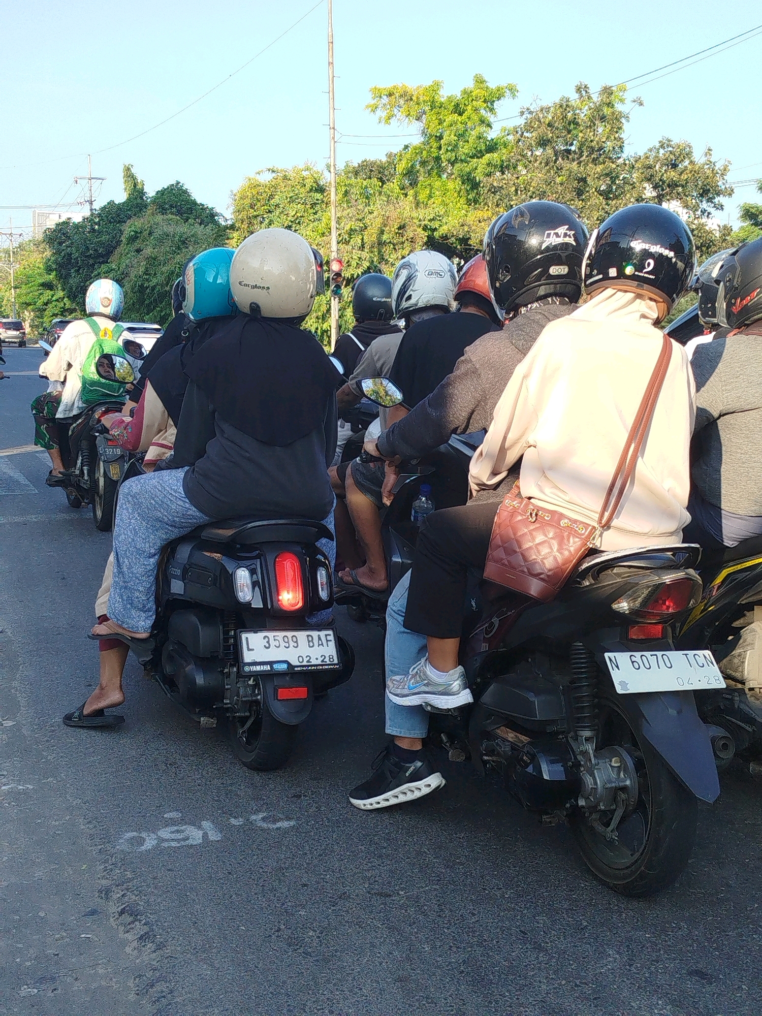 The image shows a traffic scene in Surabaya, Indonesia, where several people are riding motorcycles. The motorcycles are stopped, suggesting a traffic light or congestion. The riders are wearing helmets, with some of them visible from behind. The environment appears to be a city street with trees and utility poles in the background. The time of day seems to be late afternoon, given the sunlight. One license plate is visible and reads "L 3599 BAF" and another reads "N 6070 TCN".