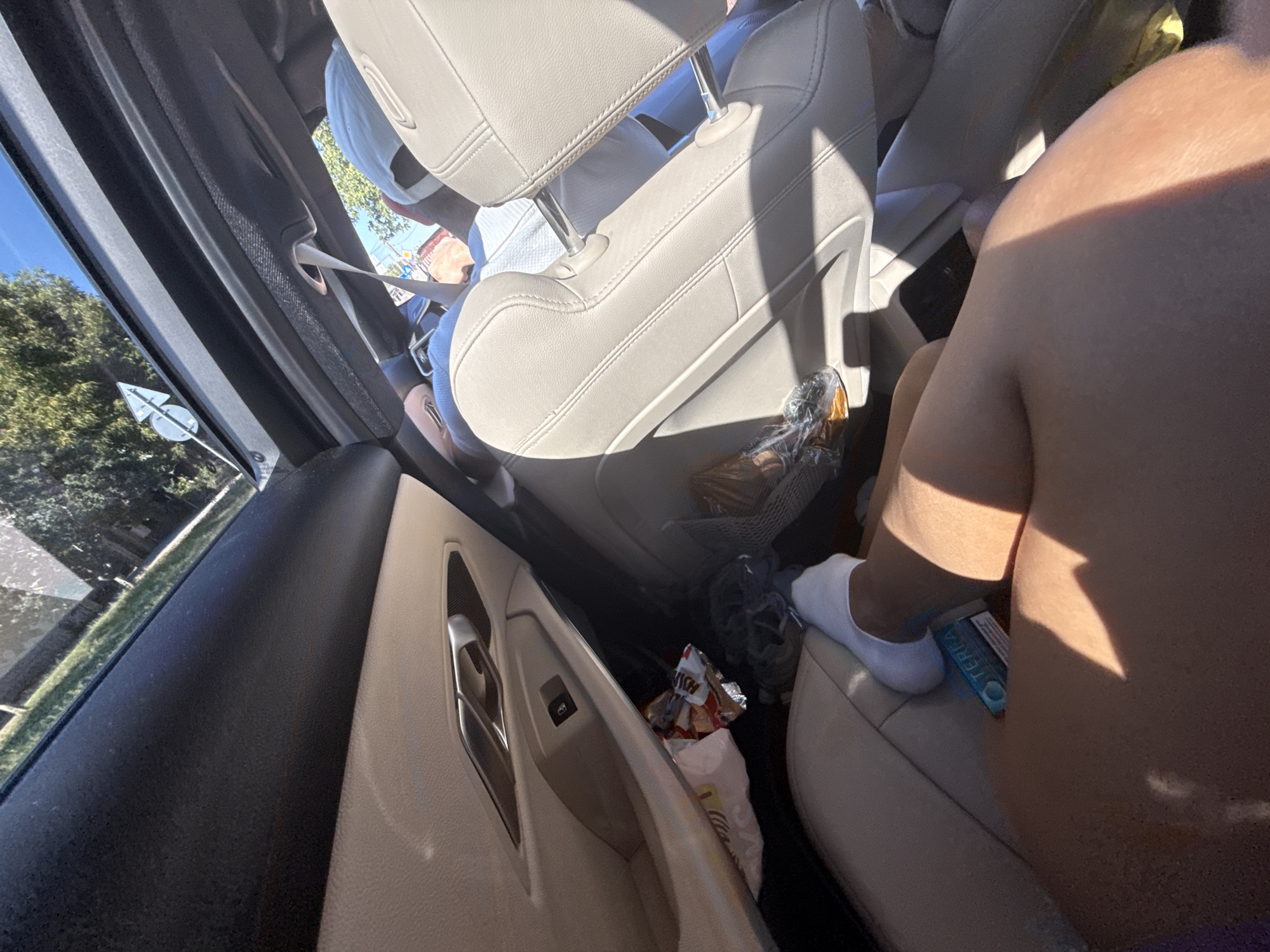 The image shows the interior of a car, possibly a convertible due to the open top. In the foreground, a person's legs are visible, with one foot wearing a white sock and resting on a blue box labeled "TEREA". The person appears to be sitting in the back seat. The interior of the car is tan and black. On the floor of the car, near the door, there are several bags of snacks, including chip bags. In the background, through the open top of the car, trees and a blue sky are visible. There are also indications of people in the front seats, one wearing a white hat. The scene suggests a casual outing or travel. It is not possible to determine the location in Simila, Romania from the image alone.