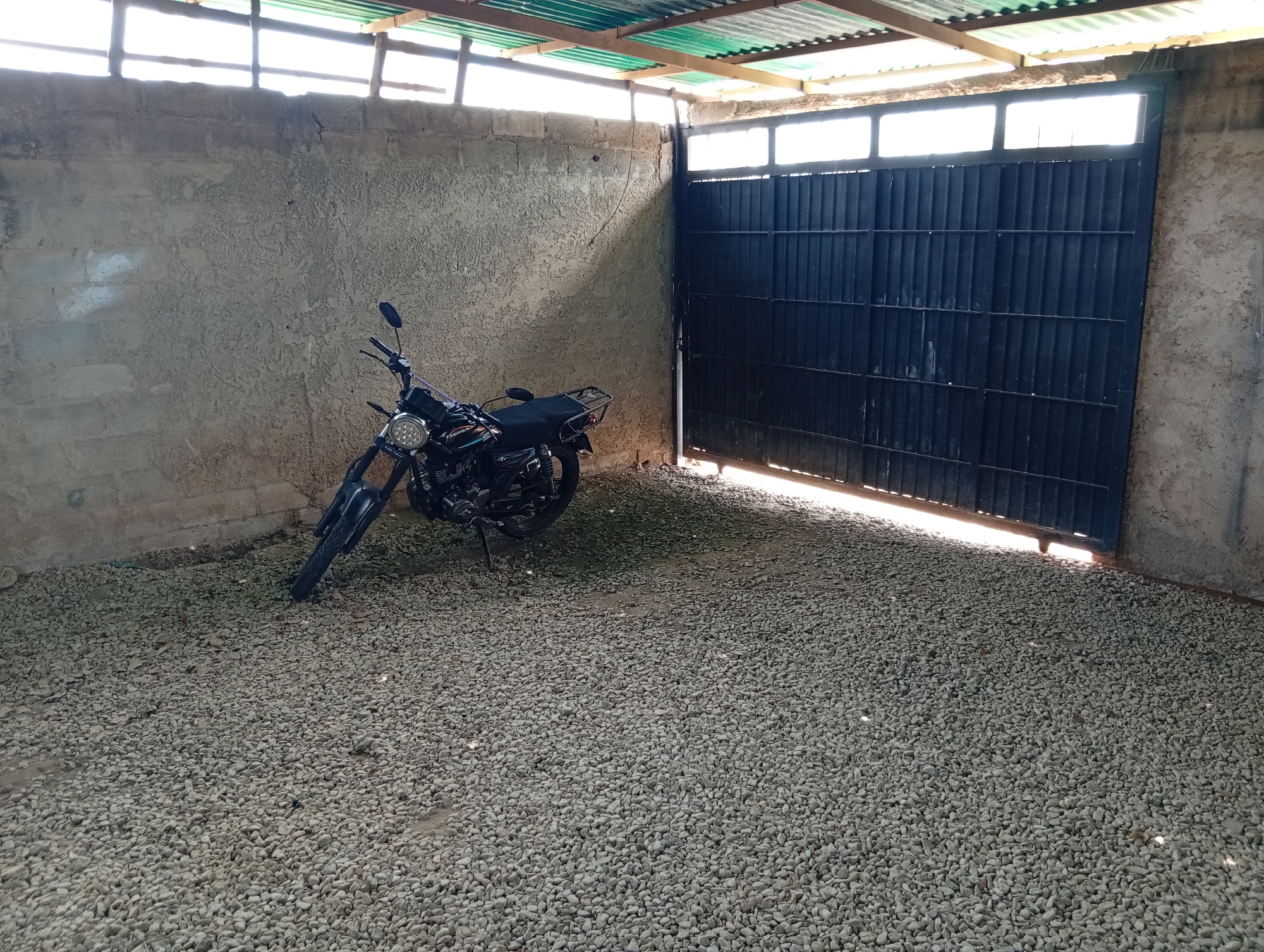 The scene is an enclosed outdoor area with a gravel ground cover. A black motorcycle is parked on the gravel against a textured wall. To the right of the motorcycle is a large, dark metal gate. The area is partially covered by a corrugated metal roof supported by wooden beams. The scene is in an area with buildings that are constructed of cinder blocks and the surroundings suggests a relatively rural or semi-rural environment. The location is Juan Griego, Venezuela. No people are visible in the frame.