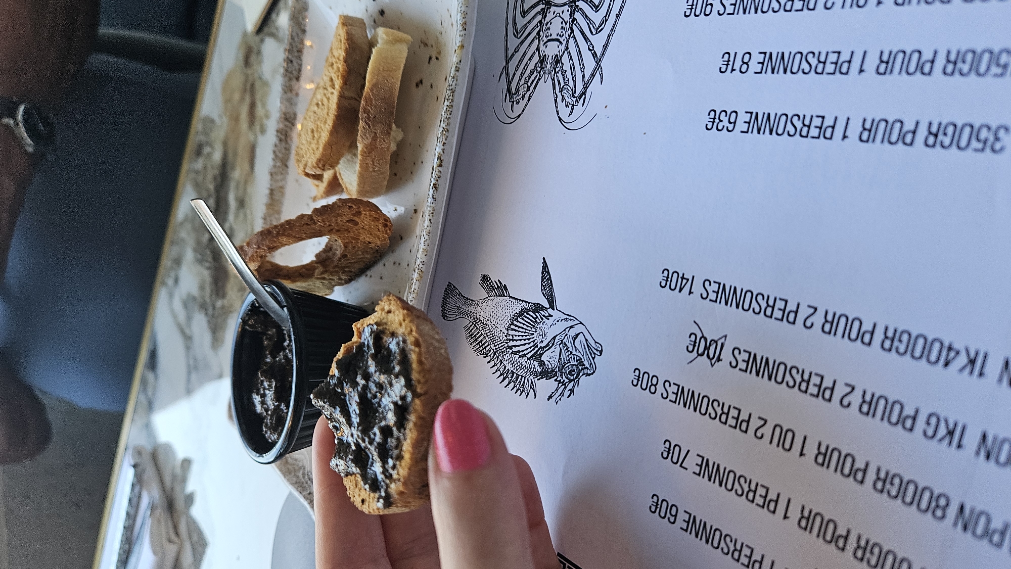 The image shows a close-up of a table setting with a hand holding a piece of bread topped with a dark spread, possibly tapenade, over a menu. To the right, on a white plate, are several slices of toasted bread and a small black ramekin containing more of the spread, with a silver spoon inside. The menu, positioned on the table, features illustrations of a fish and a lobster, and text in French indicating prices for different quantities of a food item per person. The text is angled, and partially obscured by the bread and ramekin. The setting appears to be outdoors, as suggested by the dappled lighting and the blurred background, possibly a restaurant terrace in L'Île-Rousse, France, given the presence of French text on the menu. The hand holding the bread has bright pink nail polish. The overall impression is of a pre-meal appetizer being served.
