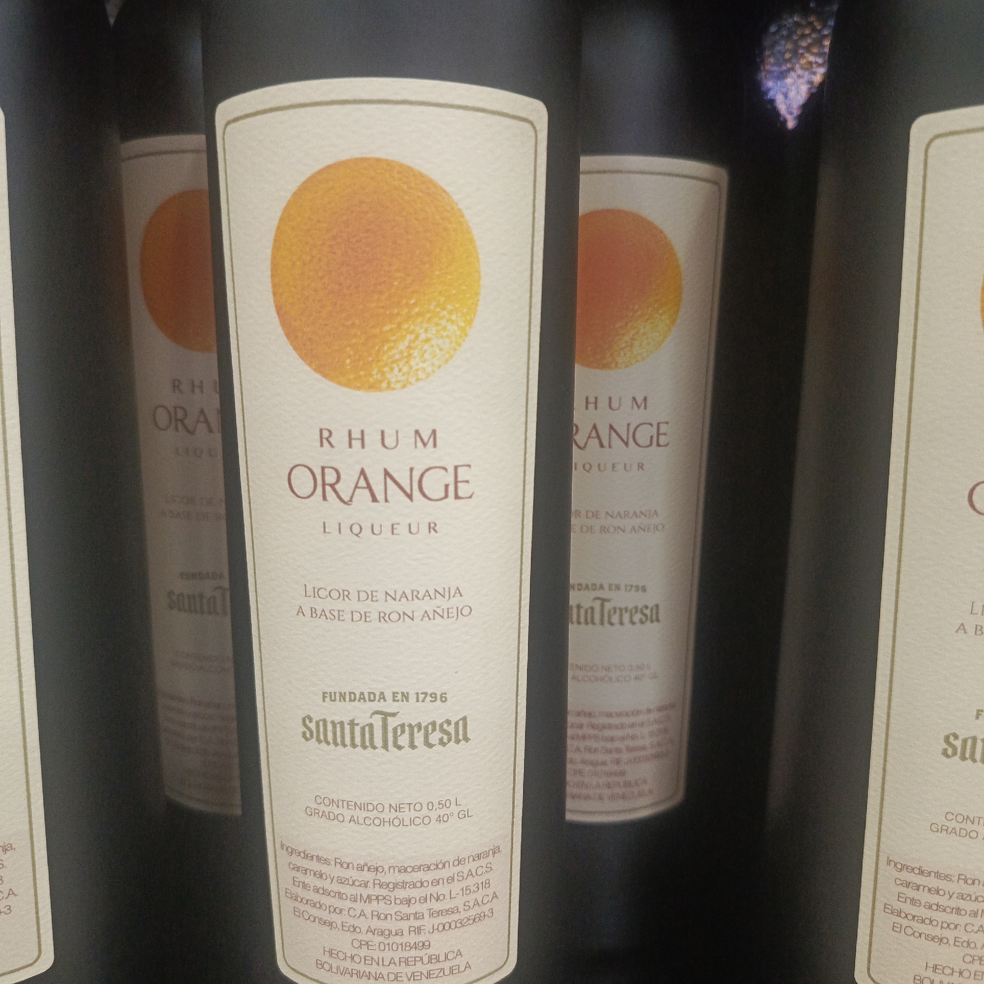 The image shows several bottles of Rhum Orange Liqueur from Santa Teresa. The bottles are arranged side-by-side, with the labels facing the camera. The central bottle's label is the most visible, displaying the brand name "Santa Teresa," "Rhum Orange Liqueur," and details about the ingredients and alcohol content. The bottles appear to be on a shelf or display, likely in a store or bar. The background is dark and out of focus, suggesting the primary focus is on the product. There are no people or specific environmental details that would allow for identification of the city of Los Teques, Venezuela.