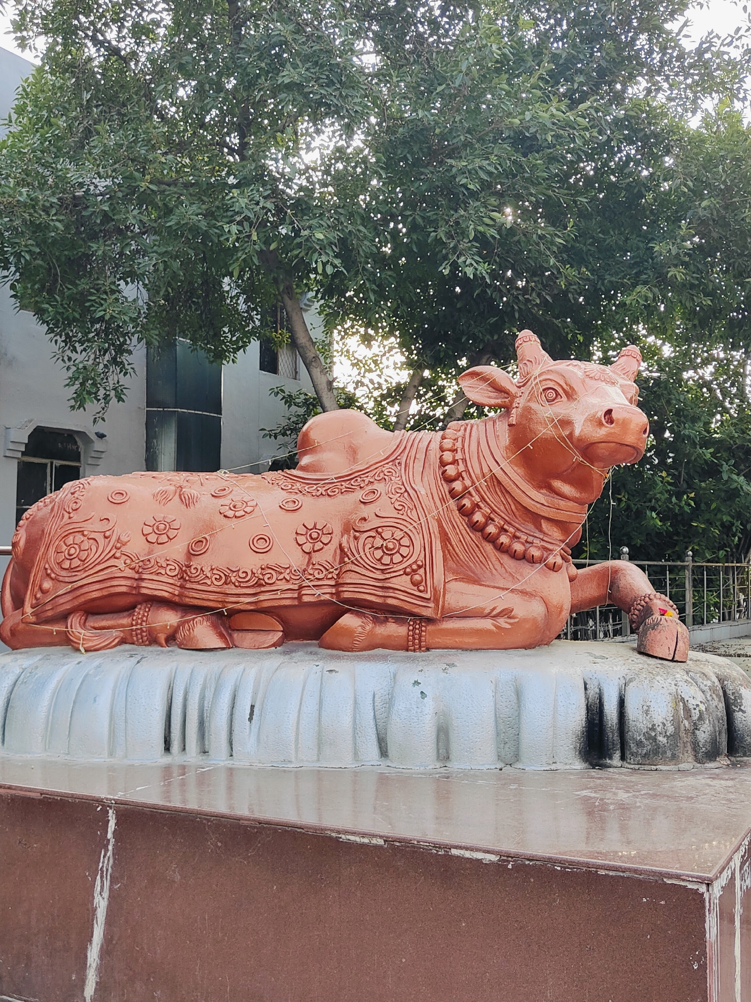 A statue of a resting bull, likely representing Nandi, the vahana of Shiva, is prominently displayed in the foreground. The statue is made of a reddish-brown material and is adorned with intricate carvings of floral patterns and decorative borders. It wears a beaded necklace and bracelets. The bull statue is positioned on a tiered, light-colored base that resembles stylized petals or waves. In the background, lush green trees with dense foliage provide a natural backdrop. Behind the trees, a modern building with windows and a grey facade is visible. The overall scene is outdoors, under what appears to be daylight, with the sun filtering through the tree canopy. The image appears to be taken at the Sheetla Mata Mandir in Gurgaon, India, a well-known religious site.