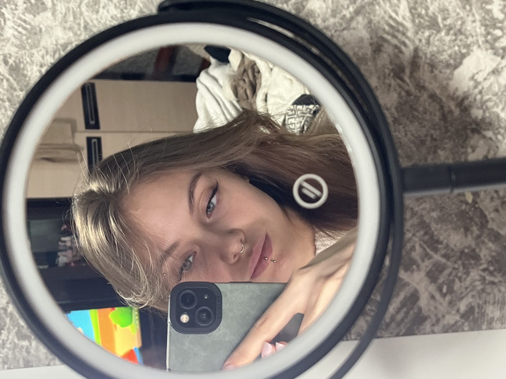 A person is reflected in a ring light mirror while holding a smartphone. The individual has fair skin, light brown hair, and facial piercings, including a nose ring, a lip piercing, and an earring. They are holding an olive green smartphone with multiple camera lenses. The ring light is white with a black frame. The background visible in the reflection shows a room with patterned wallpaper, a partially visible dresser, and some miscellaneous items. There is no definitive information in the image to confirm the location in Фоки, Russia.