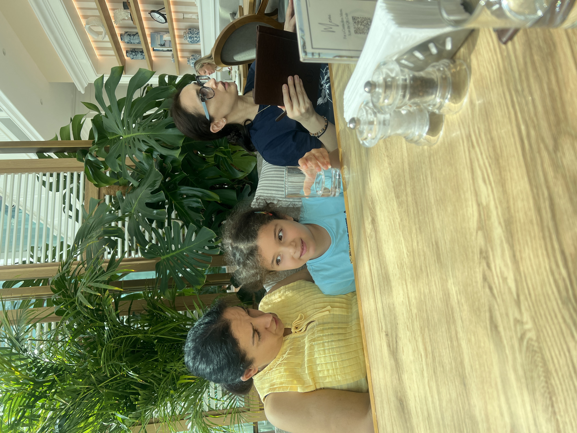 This image captures three females seated at a light wooden table in what appears to be a bright, modern restaurant or cafe in Dubai, United Arab Emirates. The setting features abundant green foliage, including large Monstera plants, contributing to a lush, indoor garden ambiance.
On the left side of the frame, a woman with dark hair, wearing a yellow textured top, is visible, looking ahead. In the center, a young girl with curly dark hair and a light blue t-shirt looks directly at the viewer with a slight smile. To the right, another woman with dark hair and glasses, wearing a dark blue top, holds a brown menu or booklet and appears to be looking up or reading it. A glass of water is placed on the table in front of the girl. On the table's right side, two clear spice grinders and a white napkin holder are present.
The background reveals decorative shelving adorned with various items like hats, small clocks, and blue and white patterned ceramic objects. A prominent sign on the table reads "menu" and instructs, "For a Contactless Menu, Scan the QR Code," accompanied by a visible QR code. The bright, natural light suggests it is daytime. The overall scene depicts a relaxed gathering or meal, with a calm and pleasant atmosphere.
