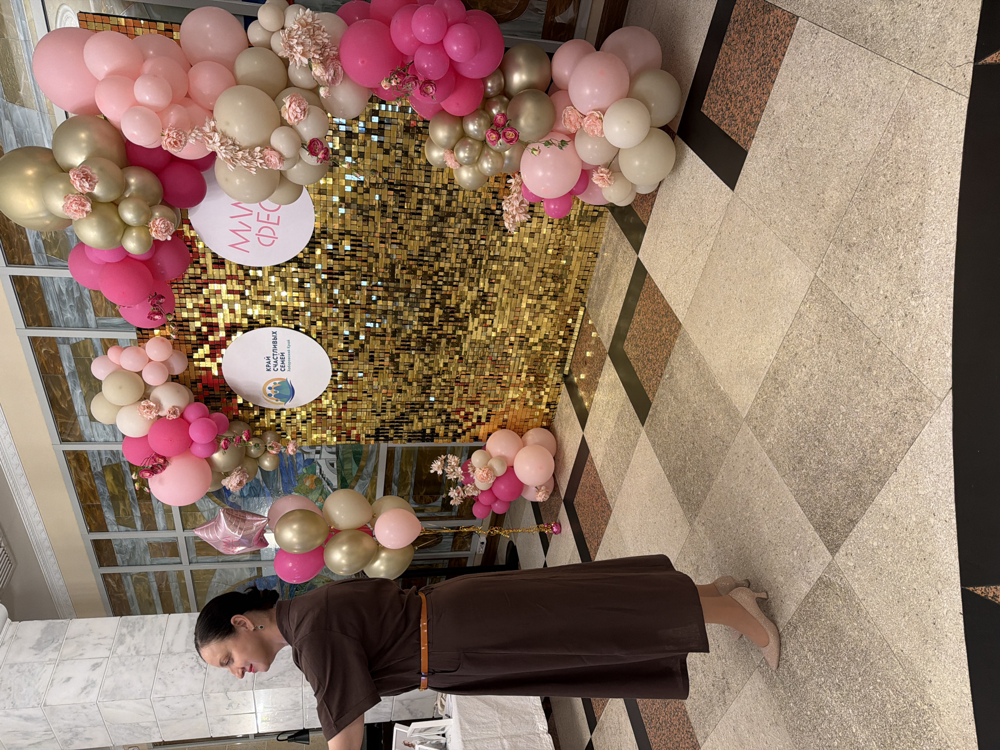 A woman in a brown dress and shimmering heels stands on a checkered floor in what appears to be a building lobby or event space in Khabarovsk, Russia. She is facing left, with her left arm extended. Behind her is a large, sparkling gold sequin backdrop arranged in a rectangular shape, acting as a focal point. An archway of balloons in shades of pink, beige, and gold frames the backdrop. Several circular white signs with text and graphics are attached to the backdrop, one of which displays the words "КРАЙ СЧАСТЛИВЫХ СЕМЕЙ" (Territory of Happy Families) and "Хабаровский край" (Khabarovsk Krai), indicating the location and theme of the event. The lighting is bright, illuminating the balloons and the woman.