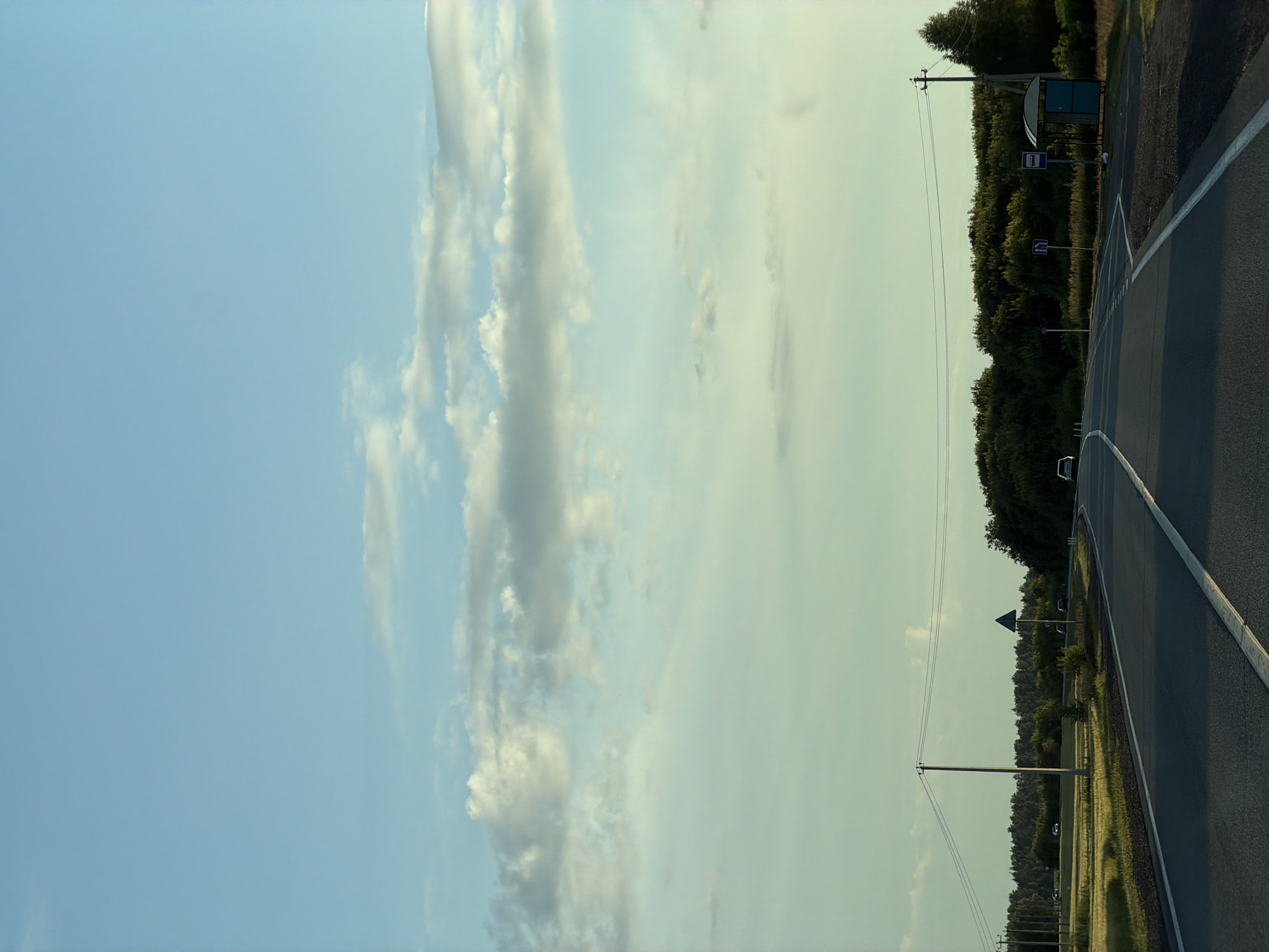 The image captures a straight, paved road in Aryshkhazda, Russia, under a bright blue sky with scattered clouds. The road is marked with white lines and appears to be a two-lane highway. On the left side of the road, a field of yellow grass stretches to a dense line of green trees in the distance. Several utility poles with wires are visible along the left side of the road. On the right side, there is a bus stop shelter with a blue roof and turquoise sides. Two blue road signs are also present near the bus stop, one indicating a pedestrian crossing and another with a blue square and white arrow. A white car is parked at the bus stop, and another car is visible in the distance on the road. The overall scene depicts a rural highway environment in Aryshkhazda during daylight hours.