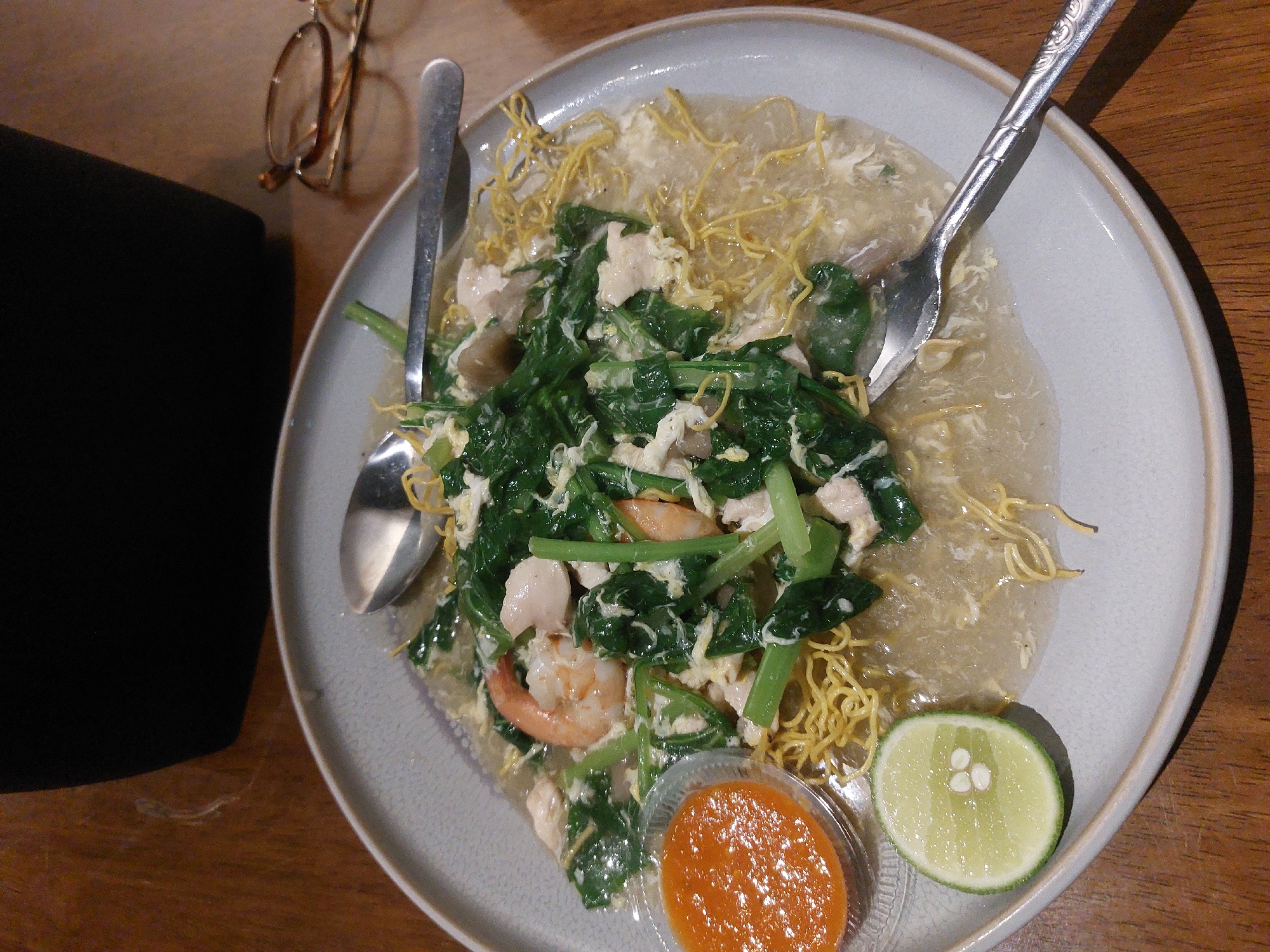 In Nunukan, Indonesia, a plate of crispy noodles with shrimp and vegetables sits on a wooden table. A small dish of chili sauce and a lime wedge are positioned to the side. Two spoons rest on the plate, one partially submerged in the dish. In the background, a pair of glasses and a dark object, possibly a bag or laptop, are visible. The lighting suggests an indoor setting, perhaps a restaurant or home.