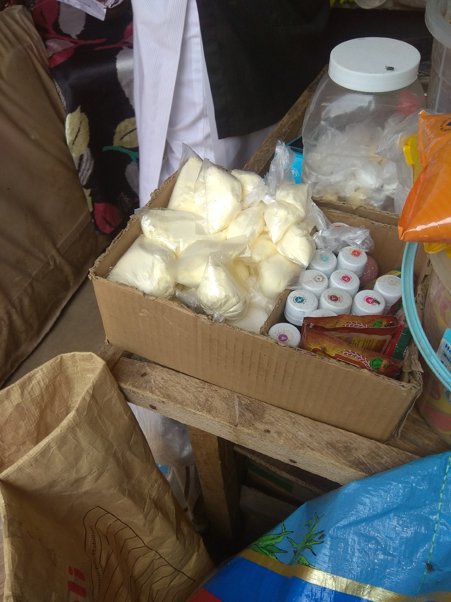 This image depicts a close-up view of a market stall in Jalingo, Nigeria. The foreground features a brown cardboard box filled with small, white, bagged powders. To the right of the box are several small, white plastic containers with colorful caps. Behind the box, a large clear plastic jar with a white lid contains more white powdery material. The background shows a patterned fabric, a white garment, and a brown paper bag. The items are arranged on wooden planks, suggesting an outdoor or open-air market setting. The overall impression is one of commercial activity involving the sale of various goods, likely food ingredients or local products.