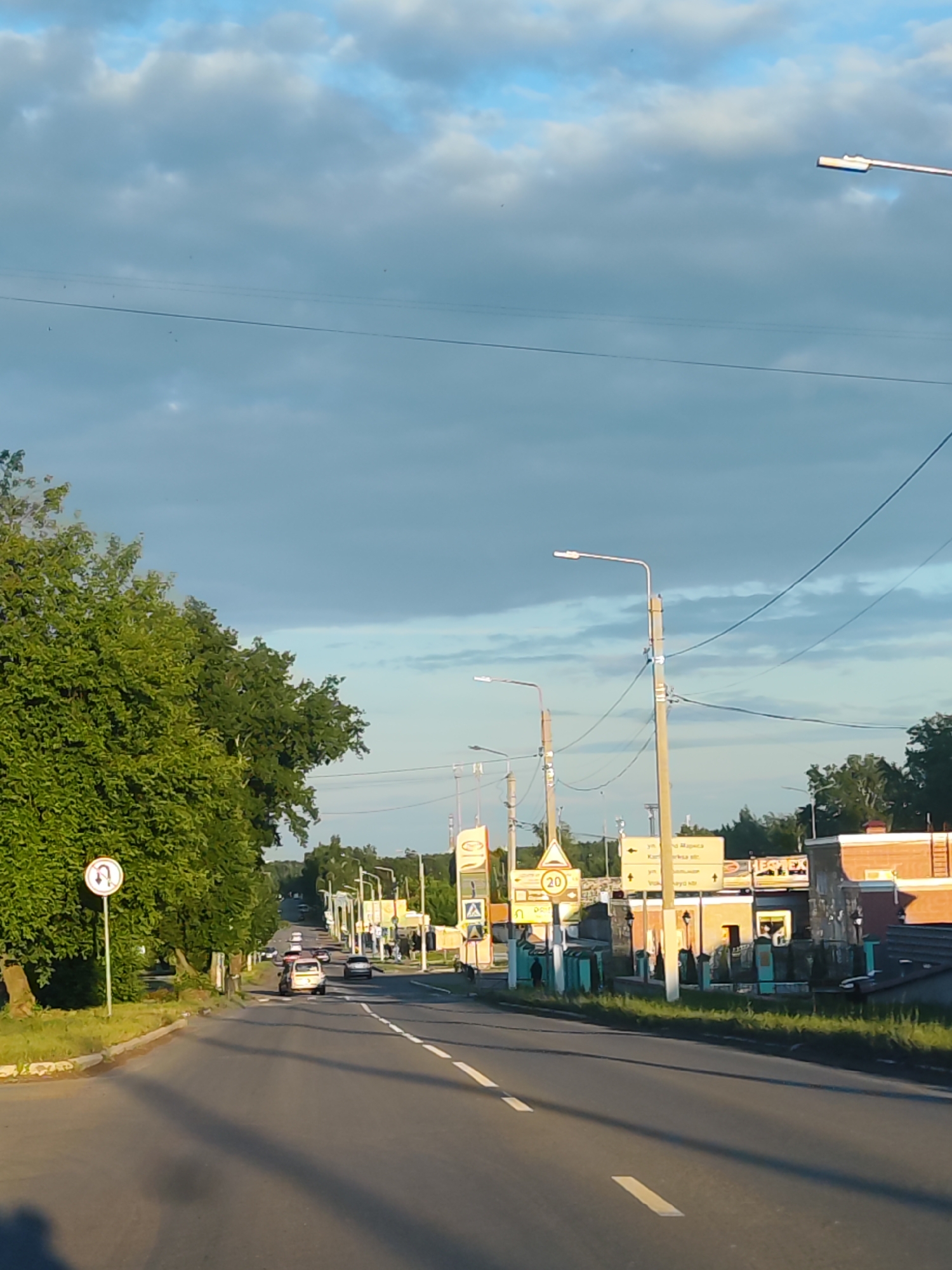 This image captures a wide-angle view of a street in Chaikovsky, Russia, during what appears to be the late afternoon or early evening. The sky is partly cloudy, with patches of blue visible among light and dark gray clouds. Sunlight casts long shadows on the road, indicating a low sun angle.
The main subject is a paved two-lane road that curves slightly to the left in the foreground and extends into the distance, appearing to go uphill. White solid and dashed lines mark the lanes. On the left side of the road, there is a dense line of green trees and grass. A "No U-turn" traffic sign (white arrow looping left with a red slash through it) is prominently displayed on the left.
Several utility poles with streetlights and overhead power lines run along both sides of the street. On the right side of the road, there are various buildings, some with brick facades, and several commercial signs. A yellow triangular sign with a black "20" indicates a speed limit of 20 km/h. Next to it, a large yellow directional sign with Cyrillic text points to different streets: "УЛ. Маркса" (Marx Street) with a left arrow, "Карповка ул." (Karpovka Street) with a straight arrow, and "УЛ. Володарского" (Volodarsky Street) with a right arrow. Other less clear commercial signs are also visible, including one with blue text.
Several cars are present on the road. In the mid-ground, a light-colored car (possibly a older model compact car or minivan) is visible, with other vehicles further down the road, including a dark sedan. The overall scene depicts a typical urban or semi-urban setting with active road traffic.