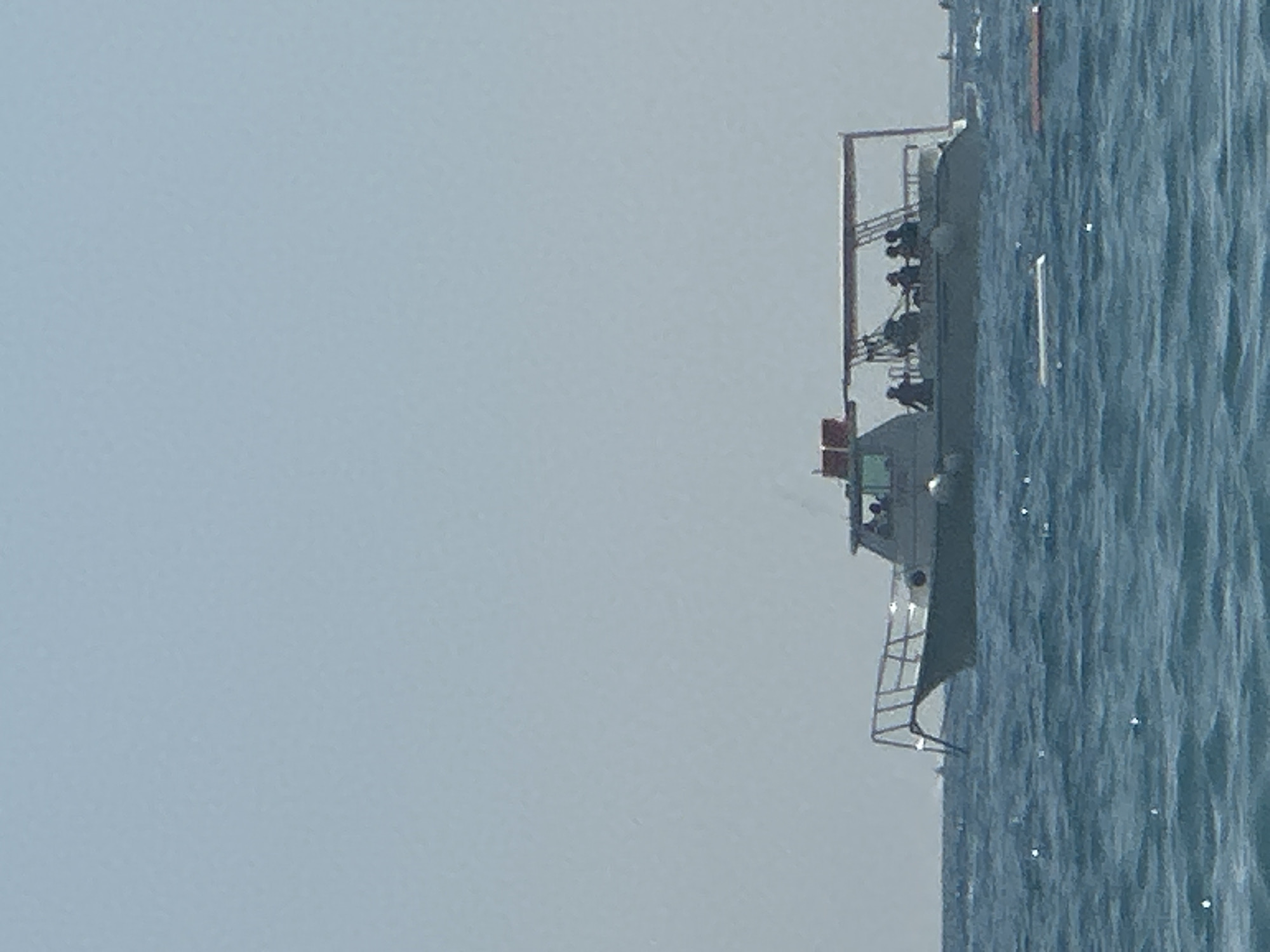 In Boreti, Montenegro, a motorboat with an open-top canopy is navigating through choppy blue waters under a clear sky. Several individuals are visible on the boat's deck. The boat has a white hull with a dark stripe running along its side and a metal railing at the bow. The boat appears to be in motion, as indicated by the waves.