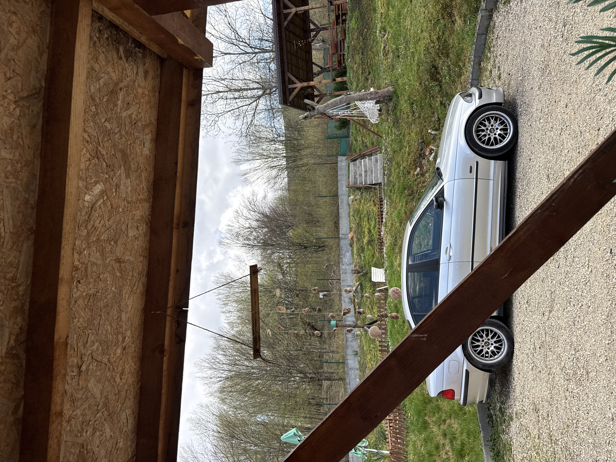 The image depicts an outdoor scene in Ribaritsa, Bulgaria, viewed from underneath a wooden structure. The foreground features the underside of this structure, constructed from OSB (Oriented Strand Board) panels and robust timber beams.
A silver sedan, appearing to be a BMW E46 model, is parked on a gravel driveway to the right of the frame. Its side faces the viewer, highlighting its multi-spoke alloy wheels. The gravel area transitions into a vibrant green lawn dotted with small white flowers, possibly daisies. A low wooden fence and a concrete retaining wall define the boundaries of the property.
In the midground and background, a rustic wooden gazebo with a dark roof stands on a grassy incline. Concrete stairs ascend towards this structure. A distinctive wind chime, fashioned from numerous seashells and suspended from a wooden beam, is visible. The landscape is dominated by deciduous trees with bare branches, suggesting either early spring or late autumn, set against a backdrop of rolling hills. The sky is partly cloudy.
No people are visible in the scene, and no discernible text is present. The time of day appears to be daytime, with natural light illuminating the area. The overall impression is a tranquil, rural or semi-rural setting.