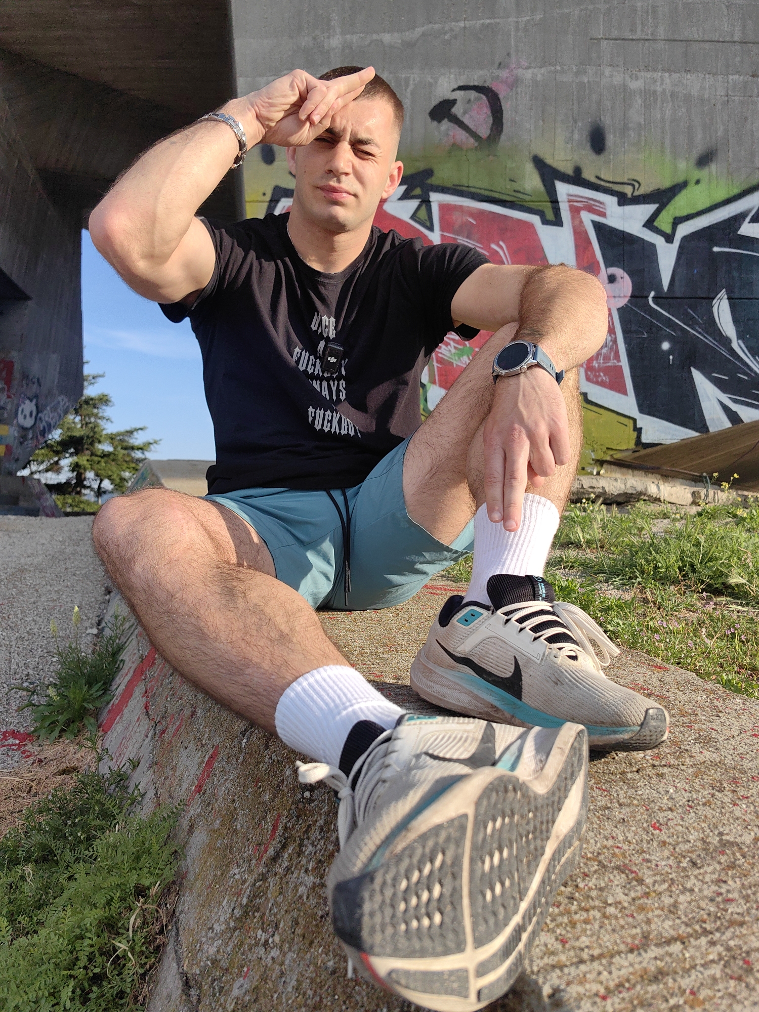 A man sits on a concrete structure, likely part of an overpass or bridge, judging by the graffiti-covered wall behind him and the glimpse of blue sky visible through an opening. He wears a black t-shirt with white lettering, light blue shorts, and white socks with running shoes. He is making a gesture with his right hand held to his forehead, as if shielding his eyes from the sun. The location of the scene is in the city of Unknown, Unknown.