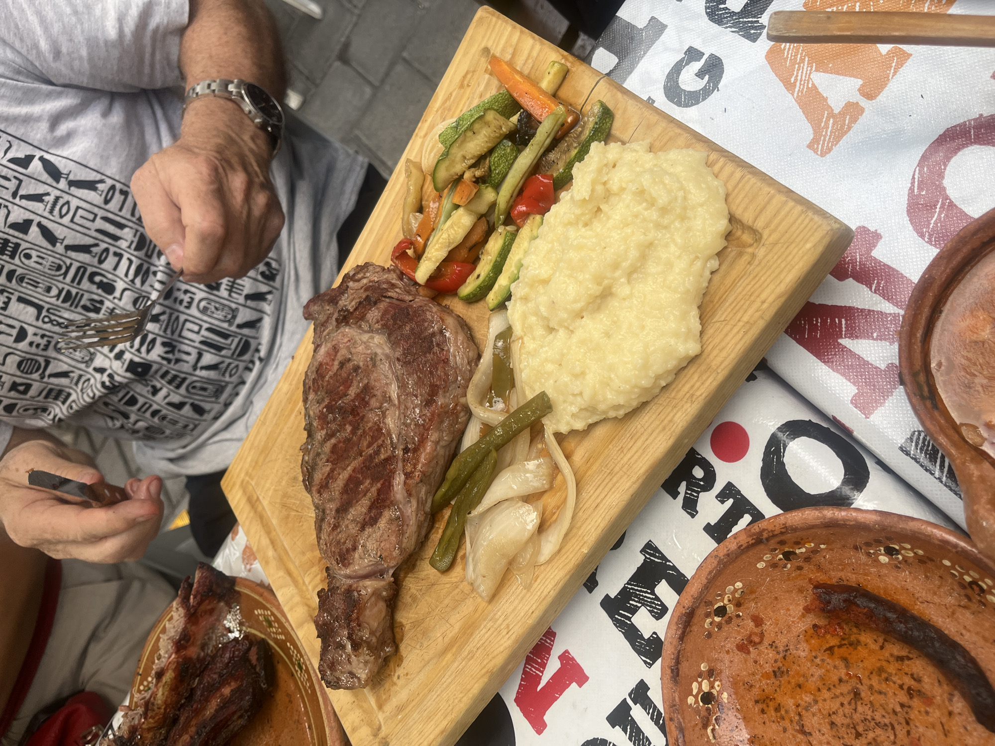 A man is seated at a table with a meal of steak, mashed potatoes, and grilled vegetables served on a wooden board. The steak is a large cut, and the vegetables include zucchini, bell peppers, and carrots. The table is covered with a patterned tablecloth. The man has a fork in one hand and a knife in the other. San Francisco Totimehuacán, Mexico, is not discernable in the image.