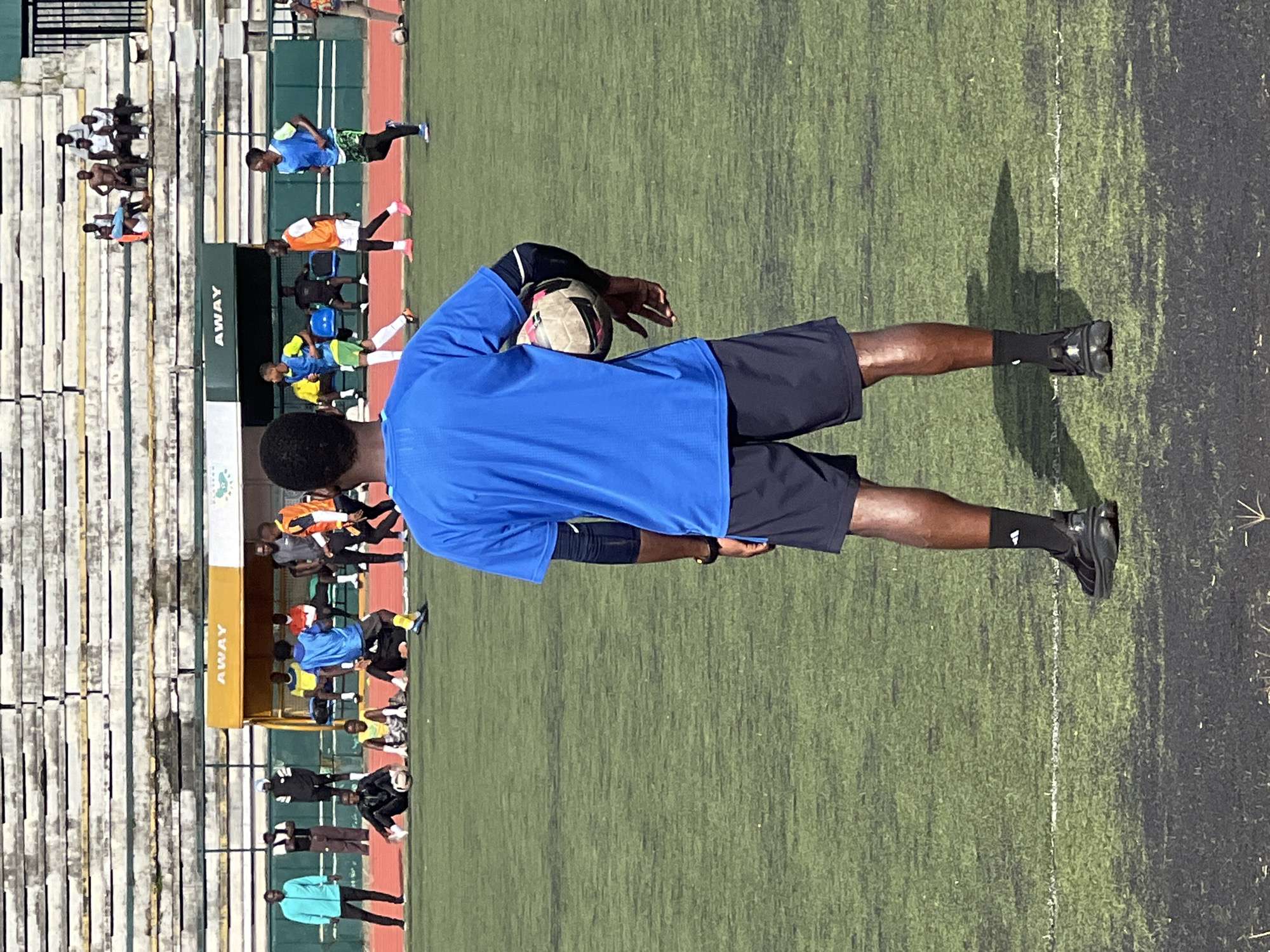 A man wearing a blue t-shirt and dark shorts stands on a soccer field, holding a soccer ball. He is facing away from the camera, looking towards a group of people on the field and in the bleachers. The field is green and artificial, with white lines marking boundaries. In the background, there are bleachers with spectators, and a building with the word "AWAY" visible on it. The scene appears to be set in a stadium in New Stadium, Nigeria, suggesting a sporting event or practice.