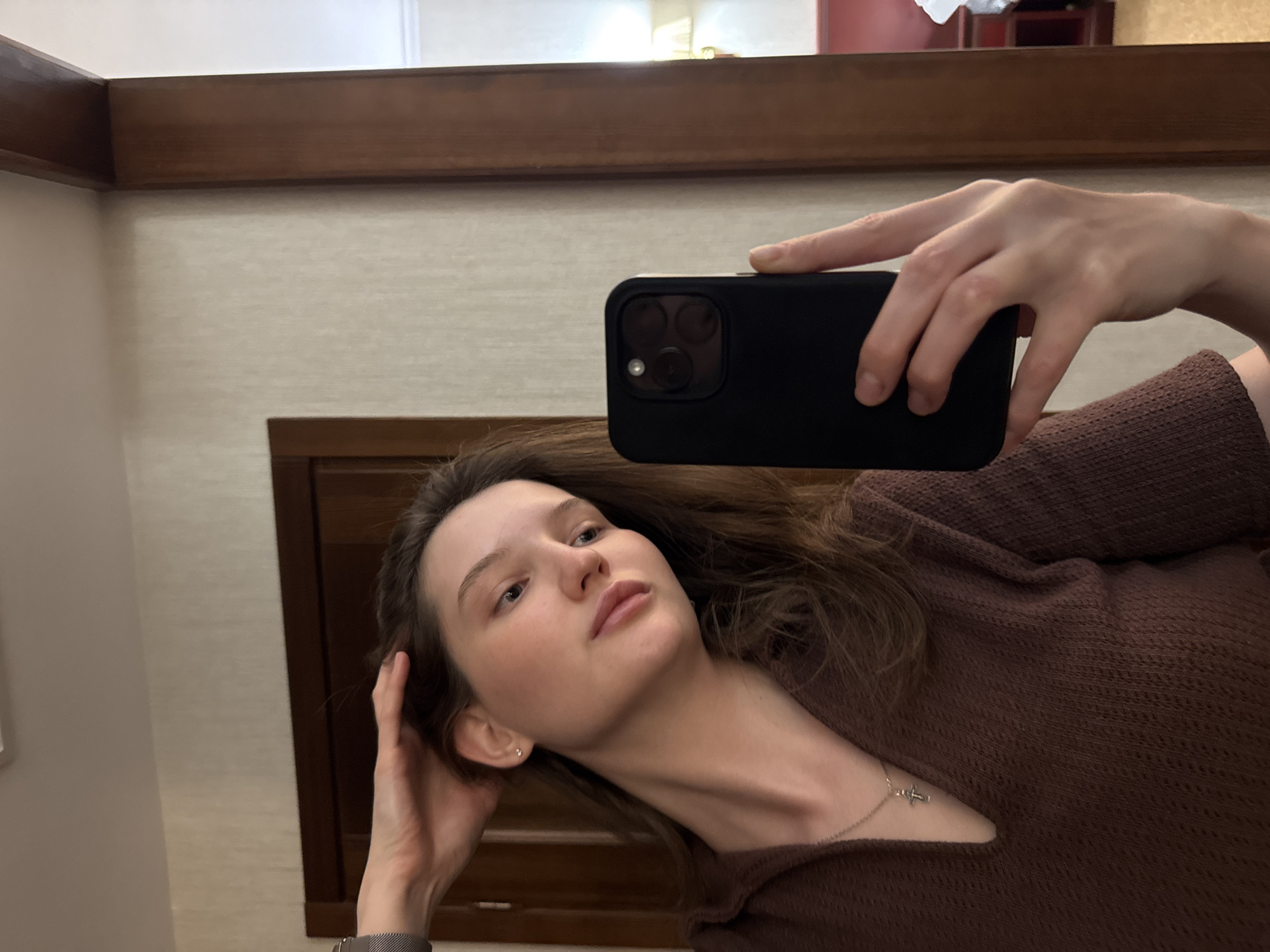 This is a portrait taken in a selfie format, featuring a young woman with long brown hair and fair skin. She is wearing a brown, textured sweater. A silver necklace with a small cross pendant is visible around her neck. Her left hand is raised, holding a black smartphone to capture the image, while her right hand is resting near her head. She appears to be indoors, with a wooden-framed mirror or wall visible in the background. The lighting suggests it could be daytime, but no specific time is discernible. The overall mood is casual and relaxed. There is no visible text in the image.