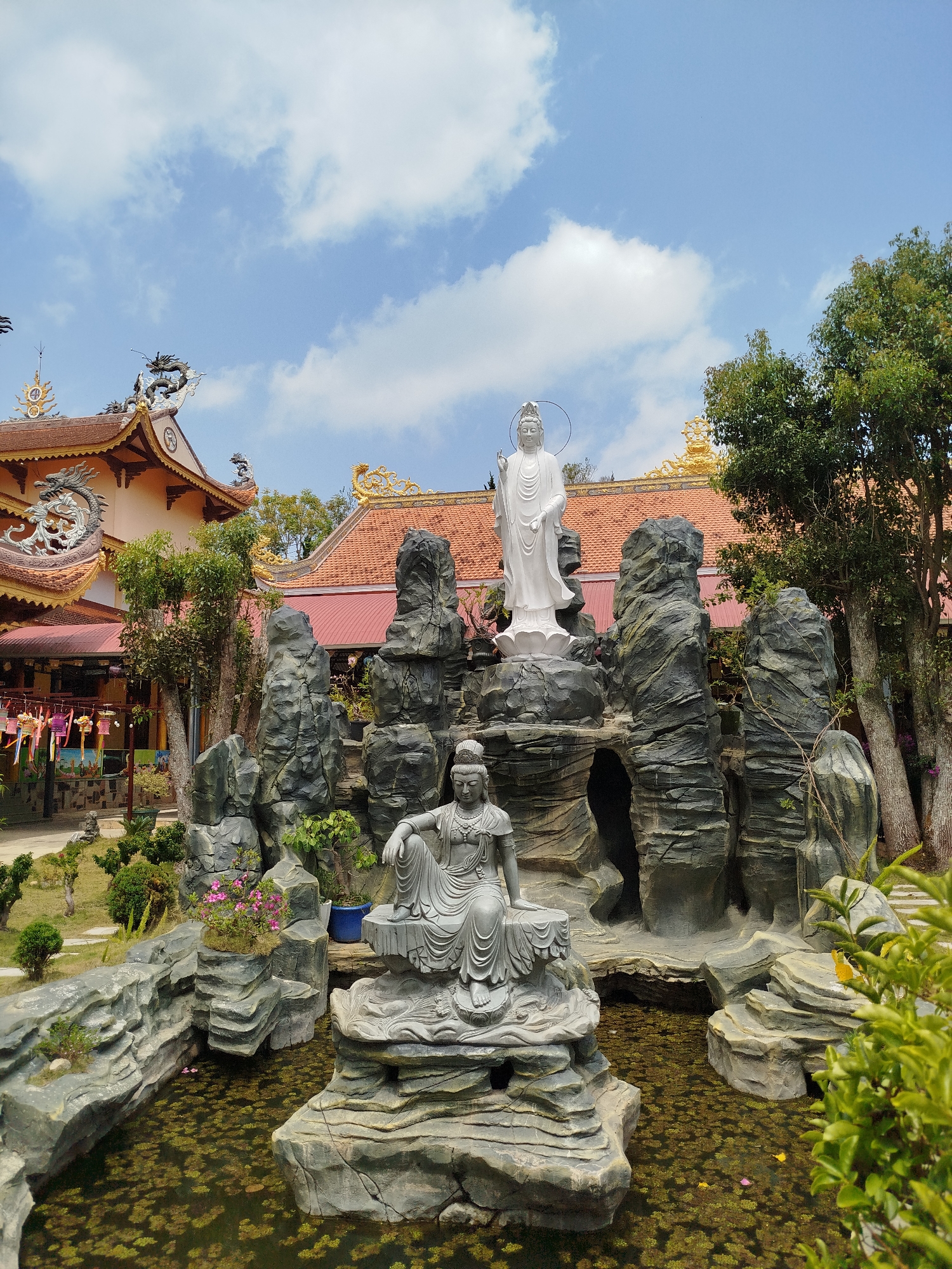 The image shows a Buddhist temple in Phát Chi, Vietnam. Two white statues are the central focus, one seated and one standing, surrounded by gray rock formations in a pond covered with green algae. The temple has a red-tiled roof with ornate golden dragon decorations. A variety of trees and other plants surround the statues and pond. The sky is blue with scattered white clouds.