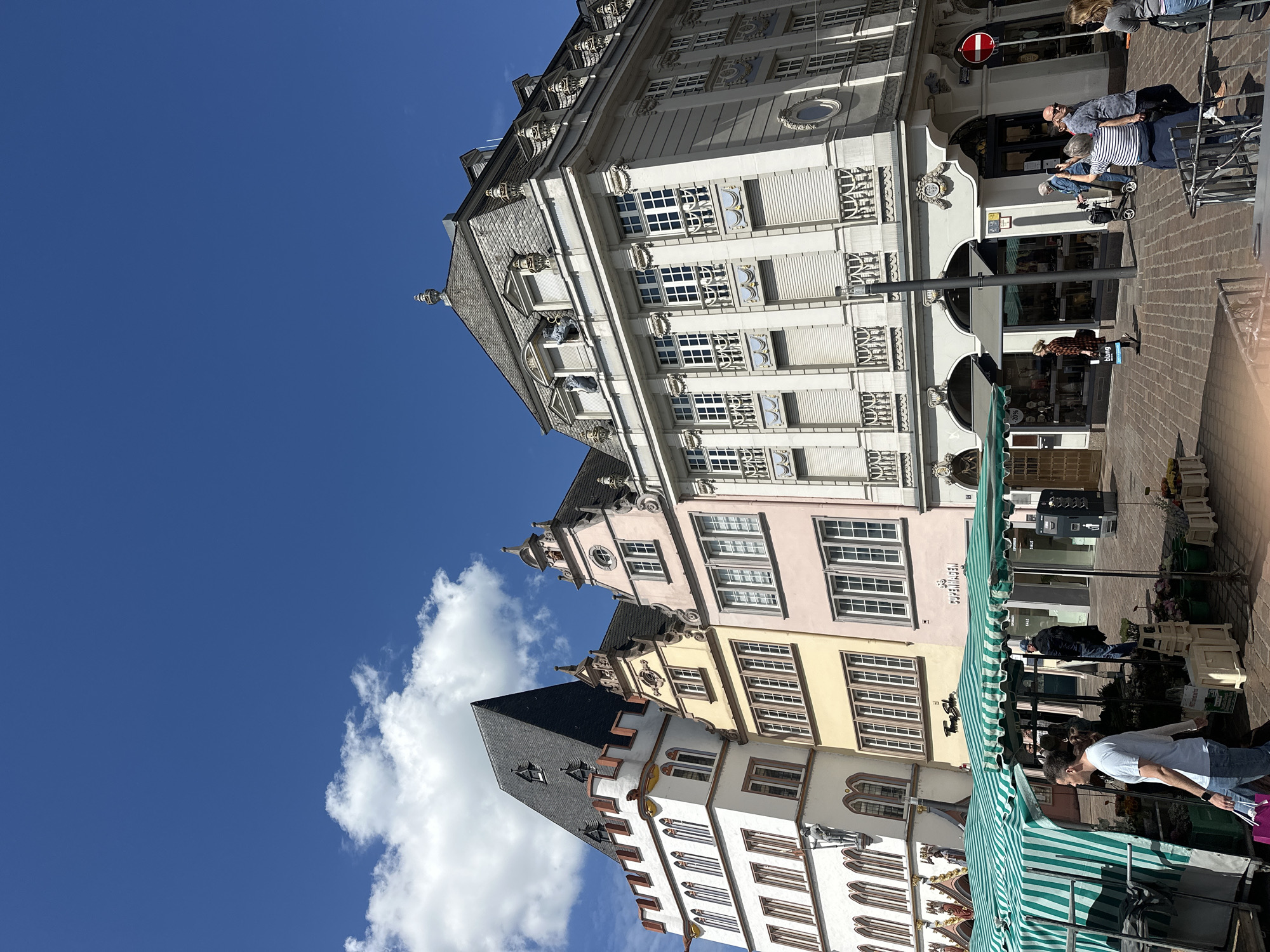 The image captures a vibrant market scene on a clear, sunny day at the Hauptmarkt (Main Market Square) in Trier, Germany. The sky, a deep blue with scattered white cumulus clouds, fills the upper half of the frame. In the foreground, several market stalls with distinctive green and white striped awnings are visible, displaying an assortment of potted plants, flowers, and other merchandise in crates and buckets.
A man in a light blue t-shirt and blue jeans walks left-to-right in the immediate foreground, carrying a purple shopping bag. Other individuals are present around the stalls, some browsing. To the left, the prominent white Dreikönigenhaus (House of the Three Kings) stands tall, featuring a steep, dark slate roof, a crenellated parapet, and multiple arched windows. A statue on a column is visible near its lower facade.
Adjacent to the Dreikönigenhaus are two multi-story gabled buildings: one with a light yellow facade and the other with a light pink facade, both featuring multiple rectangular windows and dark roofs with dormers. A shop sign reading "COPENHAGEN" is affixed to the pink building. On the far right, a grander, light-colored classical building occupies a corner, characterized by an ornate mansard roof with multiple decorative dormer windows, a richly embellished facade with pilasters, decorative balconies, and ornate window surrounds.
The ground is paved with patterned cobblestones or pavers. Numerous pedestrians are distributed across the scene, walking, standing in groups, and interacting. Among them, a woman in a striped shirt is seen raising a phone as if taking a photograph, and an elderly person with a walker is visible. A black street lamp post and a red circular "No Entry" sign are present on the right side of the frame.