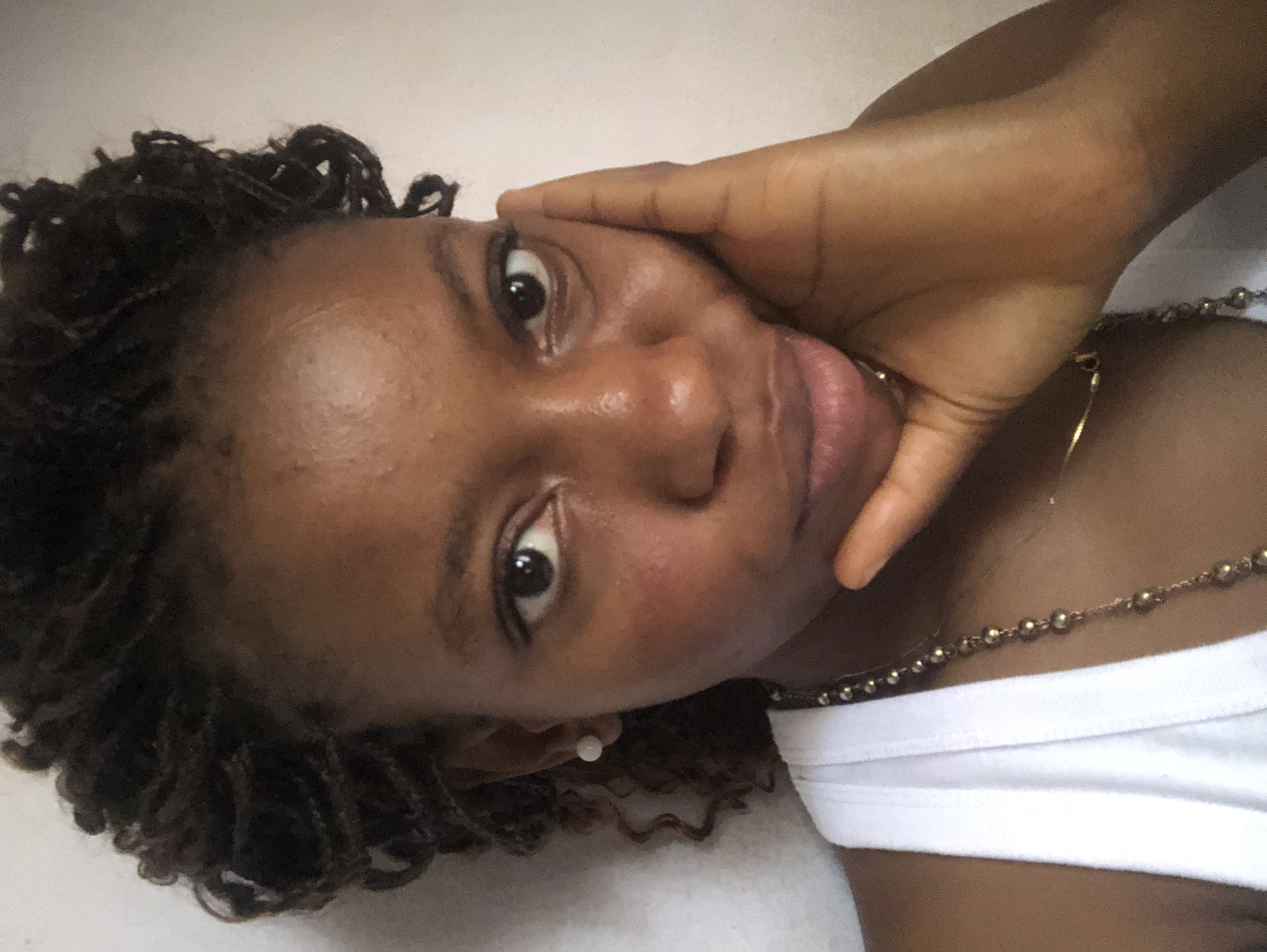 A close-up, rotated image shows a Black woman with dark brown coiled or twisted hair. She has a fair complexion, visible pores, and light brown eyes. Her right hand is positioned on her face near her temple. She wears a white tank top. A beaded necklace is visible on her neck. The background is a plain light beige or white. There are no contextual cues within the image to indicate a specific location in Kashere, Nigeria.