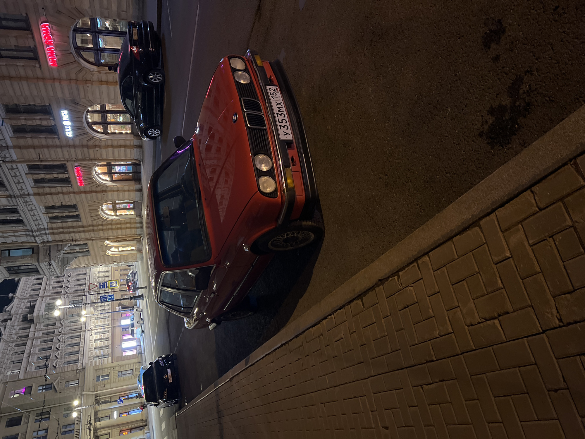The image shows a street scene in Saint Petersburg, Russia at night. An orange BMW E30 is parked on the right side of the street, and a black sedan is parked ahead of it. Buildings with illuminated windows and neon signs line the left side of the street. The license plate on the BMW reads "y353MX 152" and the plate on the black sedan reads "E996BA 98". The street is paved with asphalt, and the sidewalk is paved with bricks in a herringbone pattern. Street lights illuminate the scene. No people are visible.