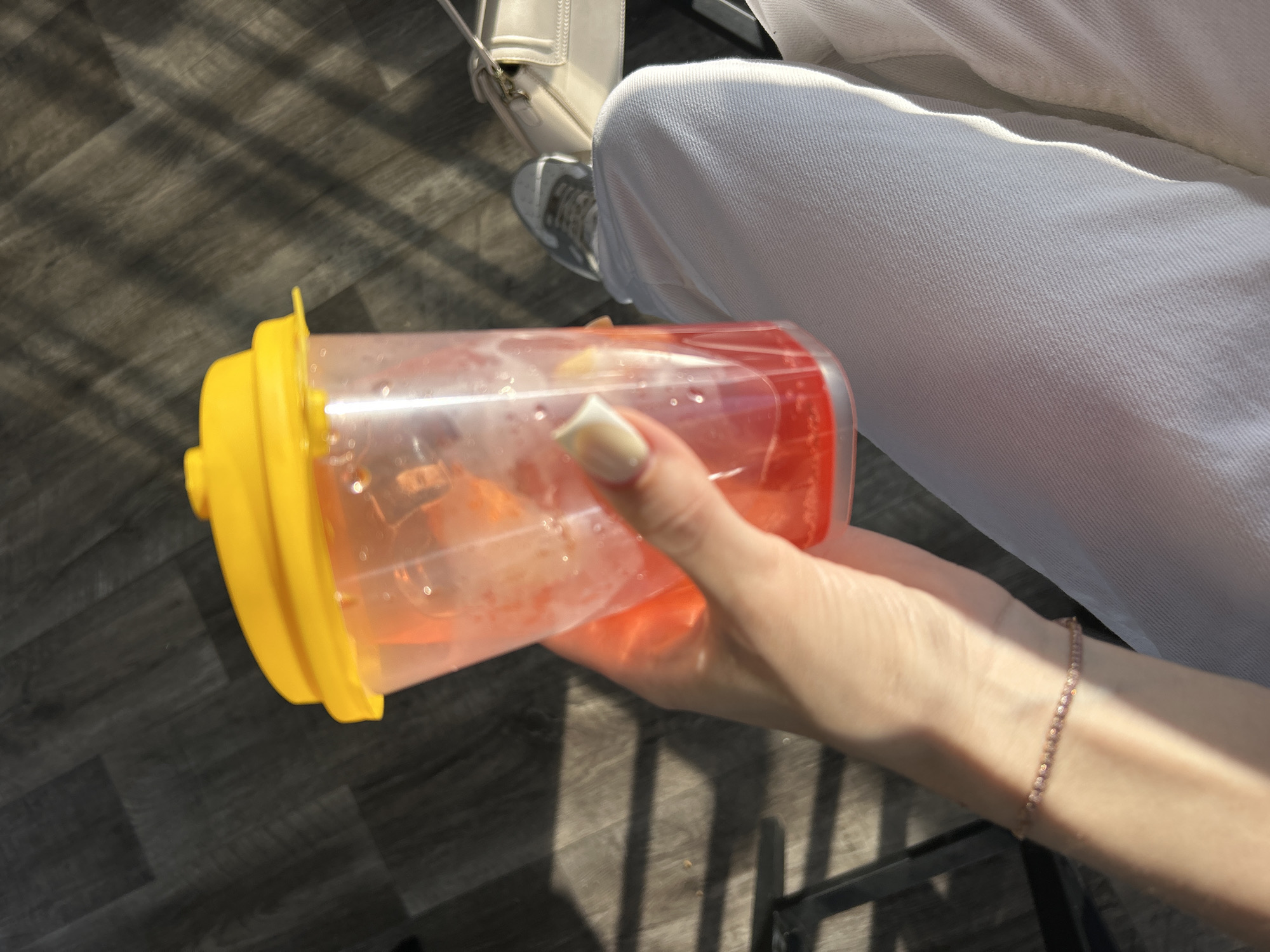 The image captures a close-up, high-angle view of a person's hand holding a drink, with parts of their leg and surroundings also visible.
**Subjects:** The primary subject is a person, though only their right hand, forearm, and parts of their lower body are in frame. Their hand, with light-colored fingernails, grips a clear plastic cup. A slender, light-colored, possibly beaded or gemstone bracelet is visible on their wrist. The person is wearing light-colored (likely white) pants, the texture of which suggests denim or a similar fabric. A white bag with a structured shape and a white shoe (possibly a sneaker with gray accents) are partially visible in the upper left background.
**Objects:** The main object is a clear plastic cup with a bright yellow lid. The cup contains a reddish-orange beverage with ice cubes and some smaller, lighter-colored pieces, possibly fruit.
**Scene and Setting:** The setting appears to be an indoor or covered outdoor area, with a dark gray floor that has a wood grain or laminate pattern. The floor is largely covered by distinct, striped shadows, suggesting sunlight is filtering through a slatted structure, like blinds or a pergola.
**Activity:** The person is simply holding the cup, possibly having a drink.
**Notable Details:**
* **Time of day:** The strong, defined shadows indicate bright, direct sunlight, suggesting it is daytime.
* **Weather:** Sunny conditions are implied by the sharp shadows.
* **Location cues:** The patterned shadows on the floor are a strong environmental cue. The dark floor and visible furniture (a dark, angular object, possibly a chair leg, in the lower right) suggest a cafe, home, or similar casual setting.
* **Visible text:** There is no visible text in the image.